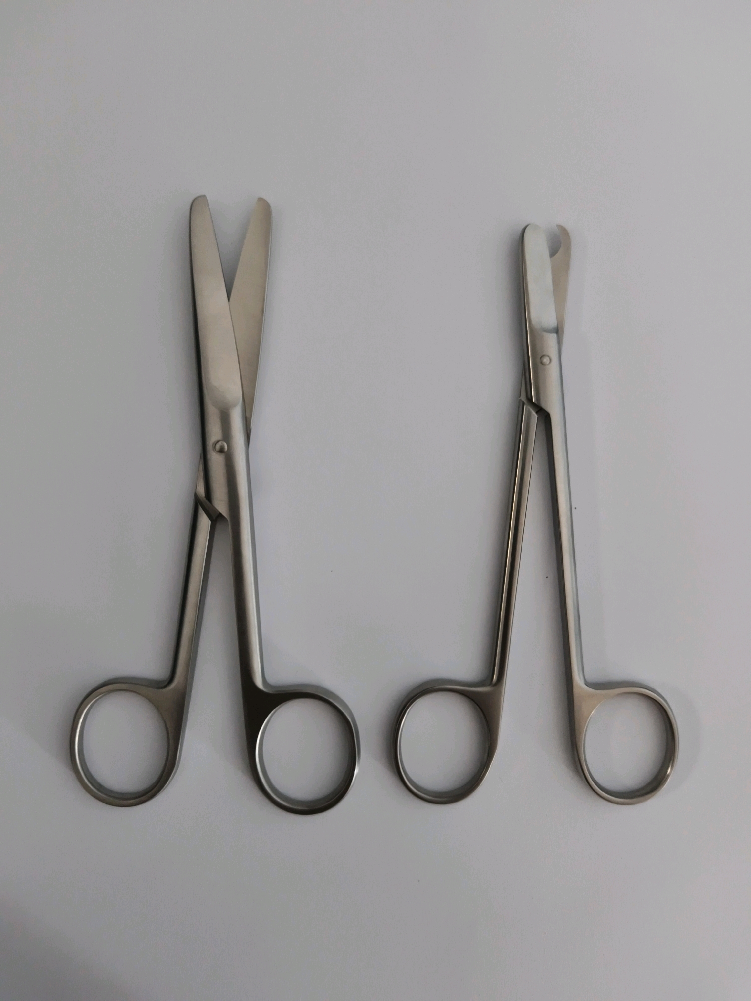 Two pairs of surgical scissors are laid out on a plain white surface. The scissors on the left have sharp, pointed blades that are slightly open. The scissors on the right have blunt, rounded tips, and their blades are closed. Both pairs of scissors are made of silver-colored metal and have finger loops for handles. The scene is devoid of any human presence or external environmental context, and therefore offers no information about the city of Tombatu Dua, Indonesia.