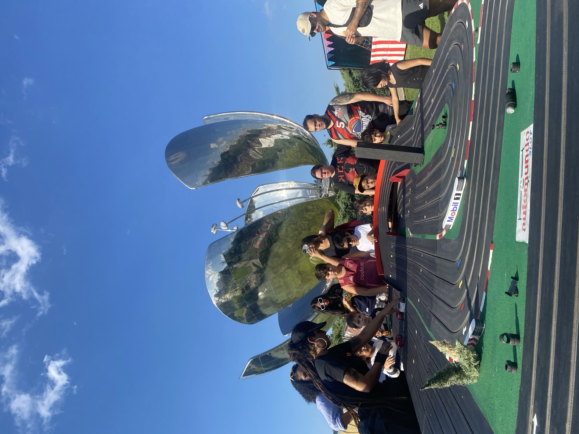 A medium, outdoor shot depicts a group of people, including children and adults, gathered around a slot car track in Buenos Aires, Argentina. In the background, under a bright blue sky, stands the Floralis Genérica sculpture, its mirrored petals reflecting the surrounding environment. The slot car track, featuring race car decals and faux greenery, appears to be the focus of the group's attention, as they observe or interact with it. Some individuals are holding phones, possibly capturing the scene or the slot car action. The weather appears sunny and clear, indicative of a pleasant outdoor setting.