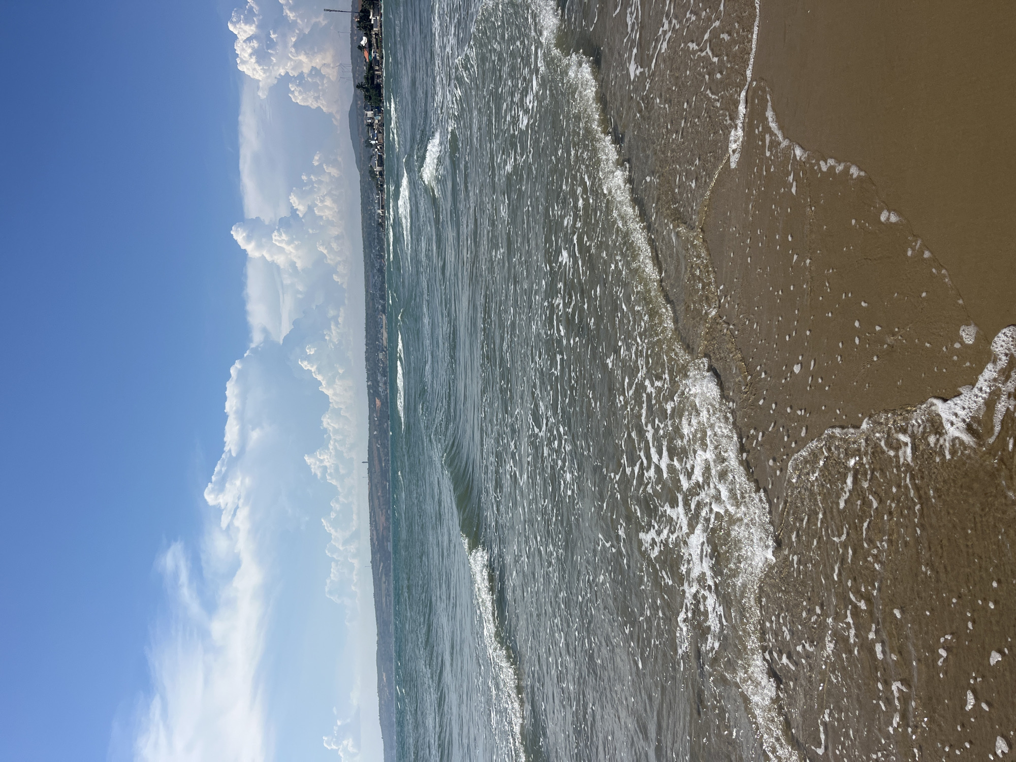 This image captures a serene beach scene in Mũi Né, Vietnam. The perspective is from the shoreline, looking out towards the ocean. The foreground is dominated by the sandy beach, with gentle waves lapping at the shore, leaving a trail of white foam and small bubbles. The water itself appears slightly murky, reflecting the sunlight and creating shimmering patterns.
In the mid-ground, the ocean stretches out, with several waves visible. The waves are not large, suggesting calm weather conditions. Beyond the water, a coastline is visible, featuring a line of buildings, likely part of the town or resort area of Mũi Né. Lush green vegetation, possibly palm trees, can be seen interspersed with the buildings, indicating a tropical setting. In the far distance, rolling hills or mountains are faintly visible, shrouded in a light haze.
The sky above is a vibrant blue, with large, fluffy white clouds scattered across it. The clouds are characteristic of a clear and sunny day. The lighting suggests it is daytime, likely late morning or early afternoon, given the brightness of the sun and the clear visibility. There is no visible text in the image. The overall mood conveyed is one of tranquility and natural beauty.