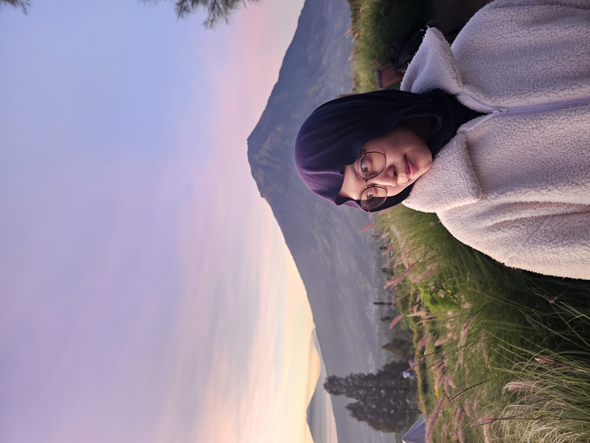 A young woman wearing a dark purple hijab and a cream-colored fuzzy jacket is captured in a close-up portrait against a backdrop of mountains and a pastel-colored sky. She has round glasses on and is looking to her left. The woman is positioned in the foreground, with tall, feathery grass in front of her. Behind her, several mountains rise, with the largest one dominating the center of the frame. The sky displays a gradient of soft pinks and purples, suggesting either sunrise or sunset. The scene appears to be an outdoor location in Tlahab, Indonesia, likely a viewpoint offering panoramic views of the mountainous landscape.