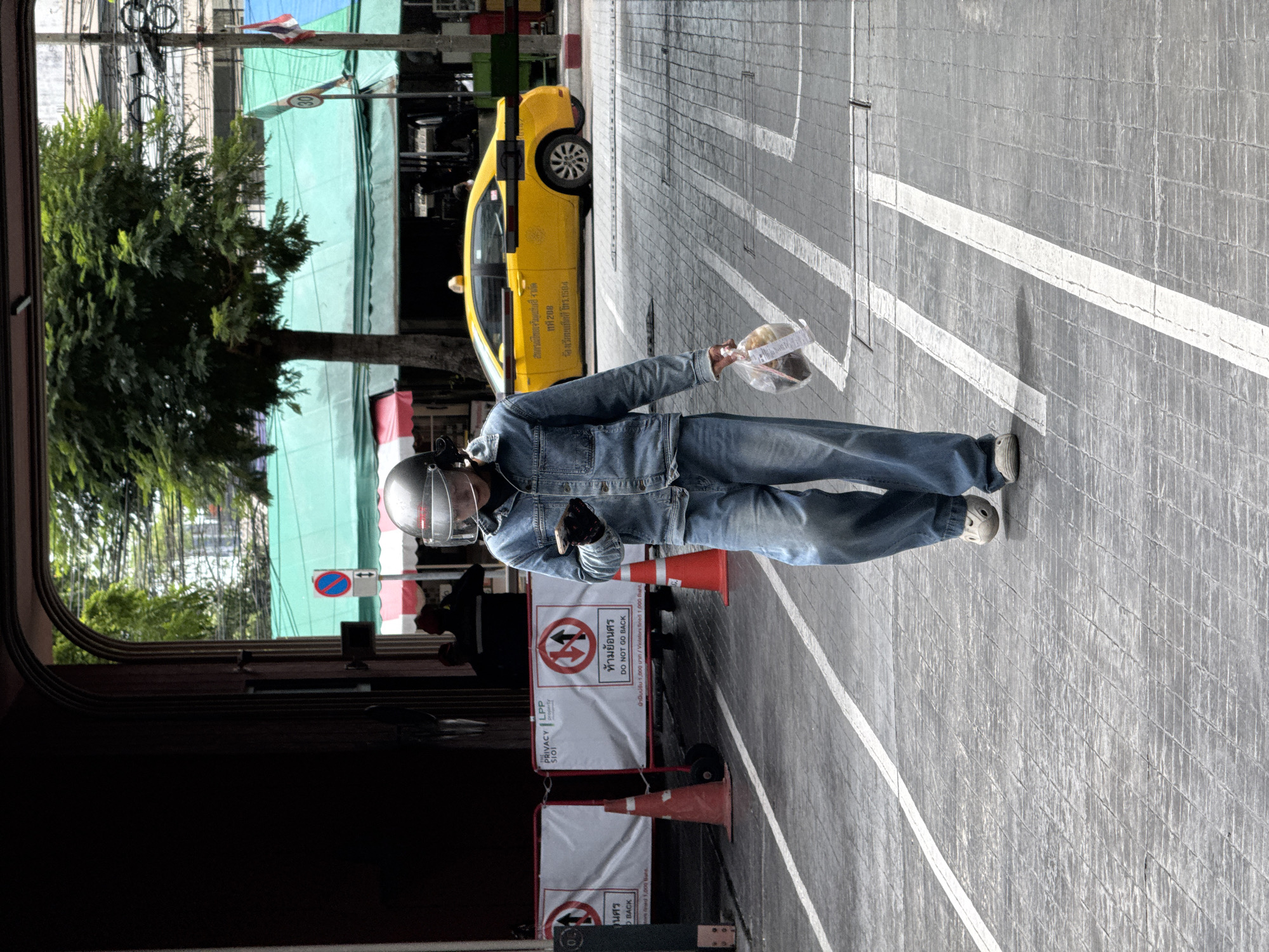 A person wearing a denim jacket, denim pants, a helmet with a clear visor, and gloves is walking on a patterned grey sidewalk in Bangkok, Thailand. The person is holding a clear plastic bag containing items in their right hand and using their left hand to interact with a smartphone. The sidewalk is marked with white lines. In the background, a yellow taxi is visible, along with a large tree and buildings with green awnings. Two orange traffic cones and several white barriers with signs are also present in the scene. The overall atmosphere suggests a street environment in a bustling city.