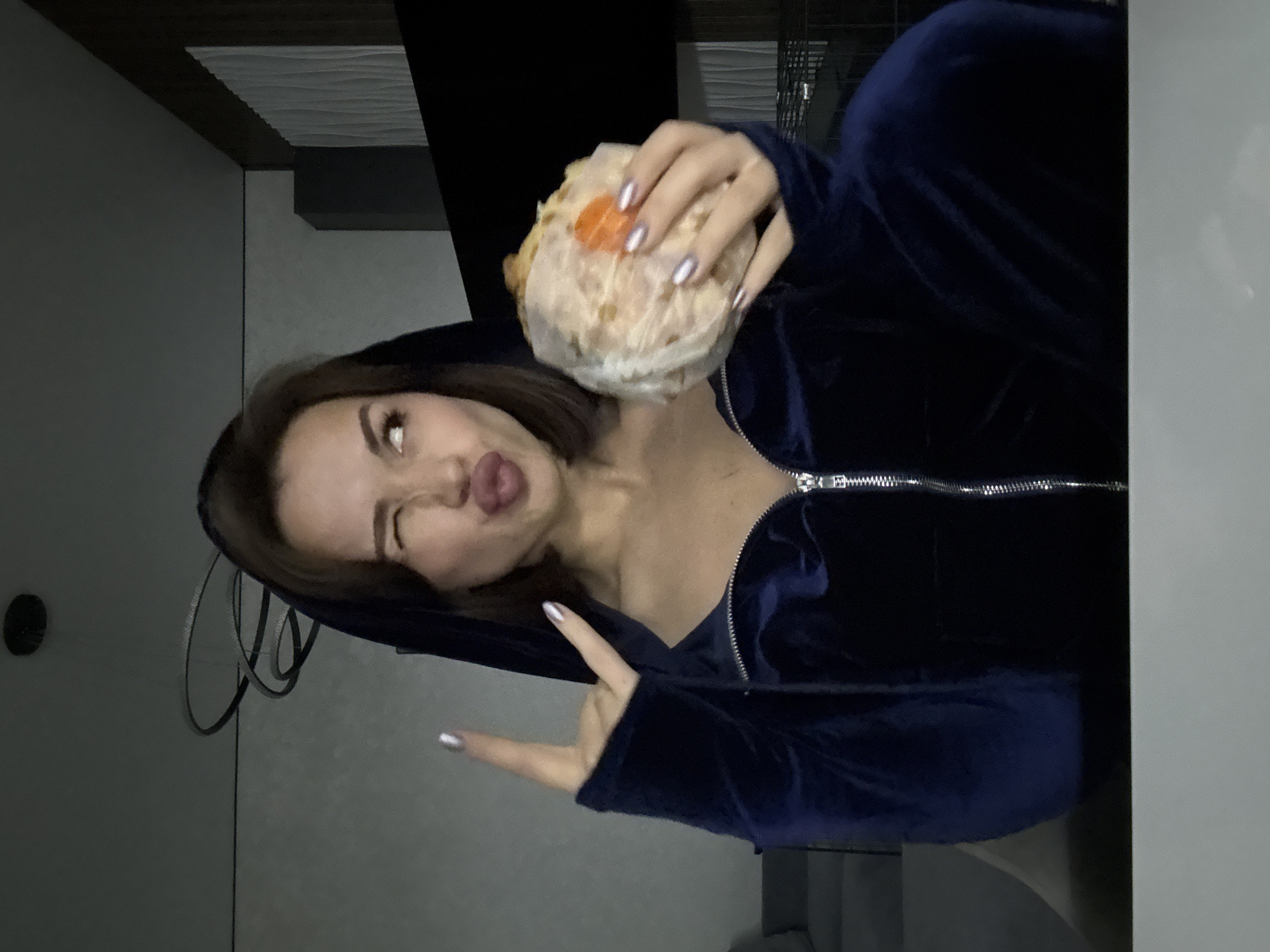 A person wearing a blue velour zip-up hoodie is holding a wrapped sandwich. The person is winking and sticking their tongue out, making a "peace" sign with their fingers. The location may be in Лобаново, Russia. Details like the background, the style of the apartment, and the person's nail polish are visible.