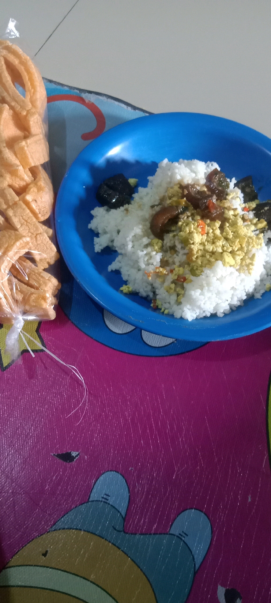 A bright blue plate holds a serving of white rice topped with scrambled eggs and pieces of what appears to be dark, stewed meat. To the left of the plate, a clear plastic bag contains an assortment of golden-brown, puffed crackers. The plate and bag rest on a vibrant pink surface adorned with a cartoonish drawing of blue and yellow characters, reminiscent of the cartoon "Dora the Explorer." This scene is consistent with a meal prepared or consumed in a household setting in Tasikmalaya, Indonesia, a city known for its culinary traditions and family-oriented culture.