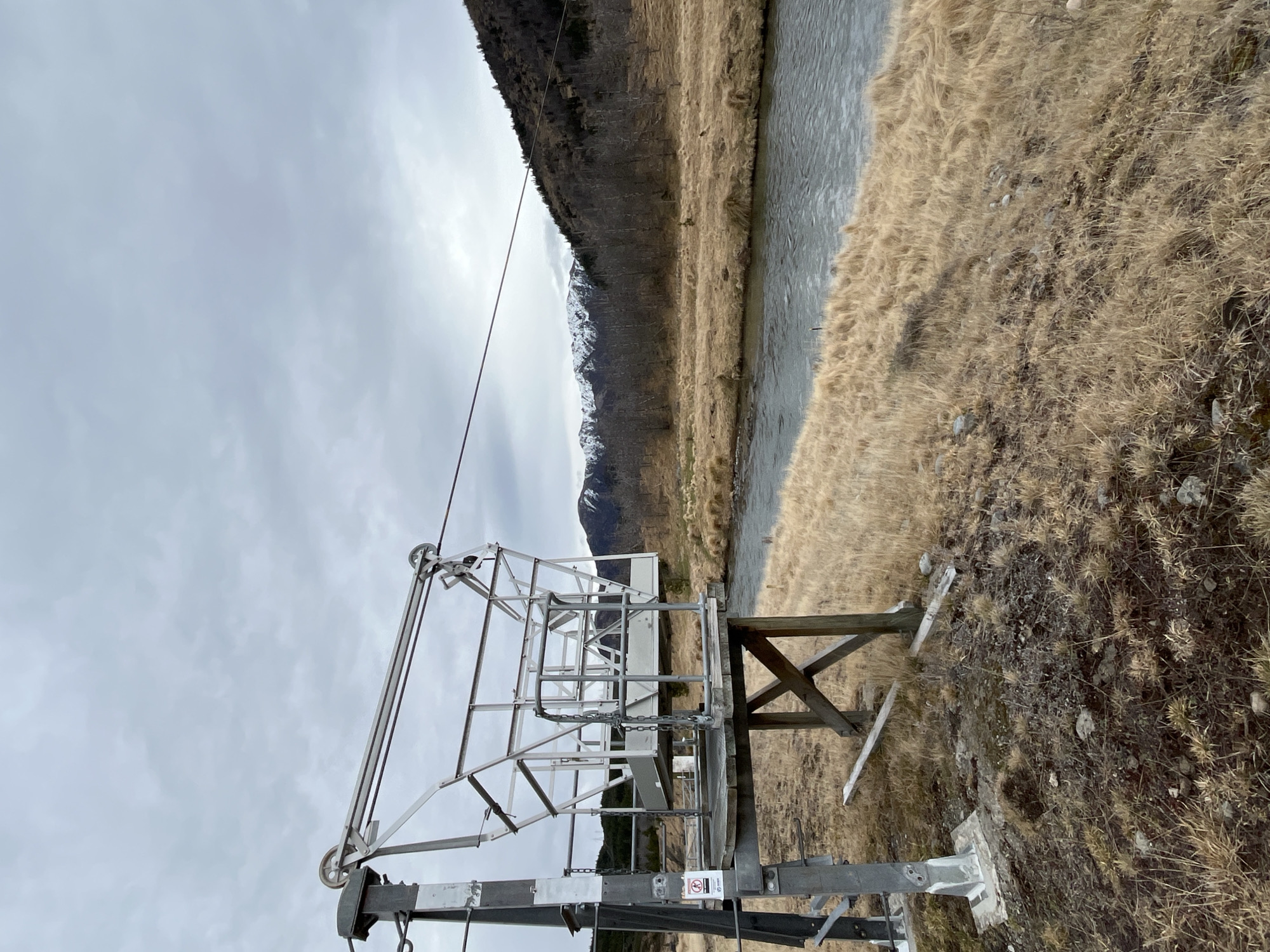 The image depicts a ski lift station in the Saint James Range of New Zealand. A metal structure with a basket-like component is situated on a wooden platform supported by an X-shaped frame. A cable extends from the structure across the frame and into the distance. A small sign with a red circle and a diagonal line is attached to the metal support post. A river flows to the right of the ski lift, bordered by dry, golden grass. Beyond the river, a treeline leads to a mountain slope covered in sparse trees and snow-capped peaks under a cloudy sky. The ground around the ski lift is rocky and covered with dry vegetation.
