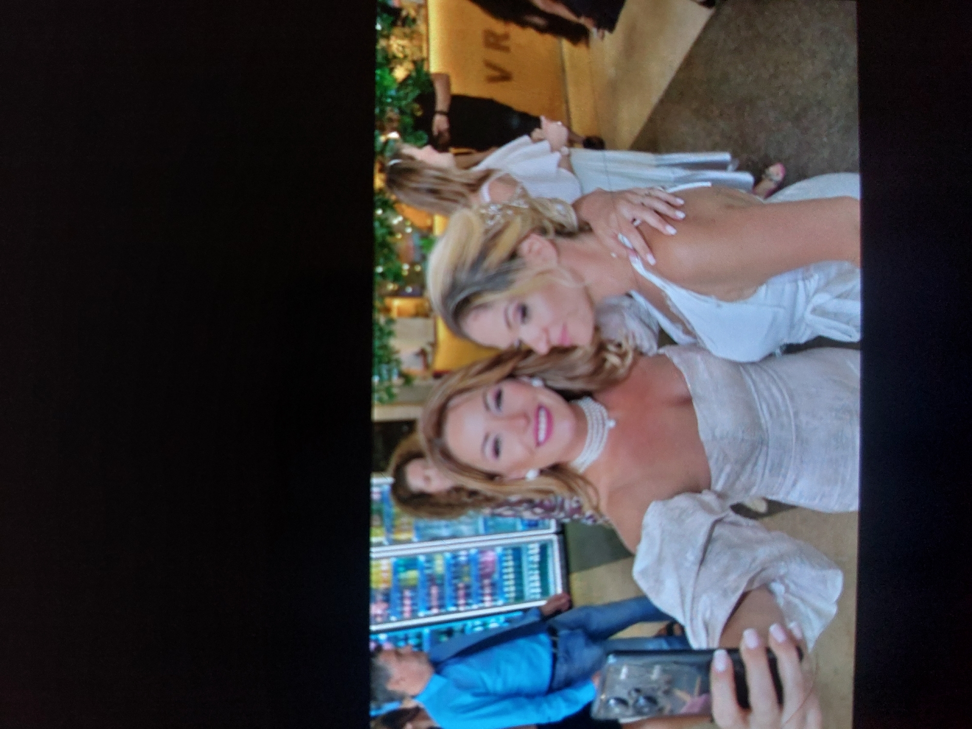 The vertically oriented image captures an indoor social gathering, likely in a restaurant, bar, or hotel lobby in Tsarevo, Bulgaria. The primary subjects are two smiling women, appearing to be friends, posing for a photograph.
The woman in the foreground has blonde hair and is wearing a light-colored, off-the-shoulder top or dress made of textured fabric. She is adorned with a pearl choker necklace and matching pearl earrings, and her lips are highlighted with bright pink lipstick. She holds a smartphone, suggesting she is taking a selfie or group photo, and her fingernails display a light, possibly ombré, manicure. Behind her, the second woman, also with blonde hair, wears a white, sleeveless top or dress. She is seen embracing the first woman, leaning in with a smile and a slight pout towards the camera.
In the background, several other people are visible, indicating a busy setting. To the left, a man in a blue shirt and jeans stands near beverage coolers stocked with drinks. Further back, a yellow wall or bar counter is prominent, with the letters "VR" faintly visible on its surface. Green decorative foliage or garlands add to the ambiance around this structure. The flooring appears to be carpeted or tiled in neutral tones. The lighting suggests an indoor evening event or a brightly lit interior. The overall atmosphere is joyful and sociable, with the women expressing happiness and camaraderie.