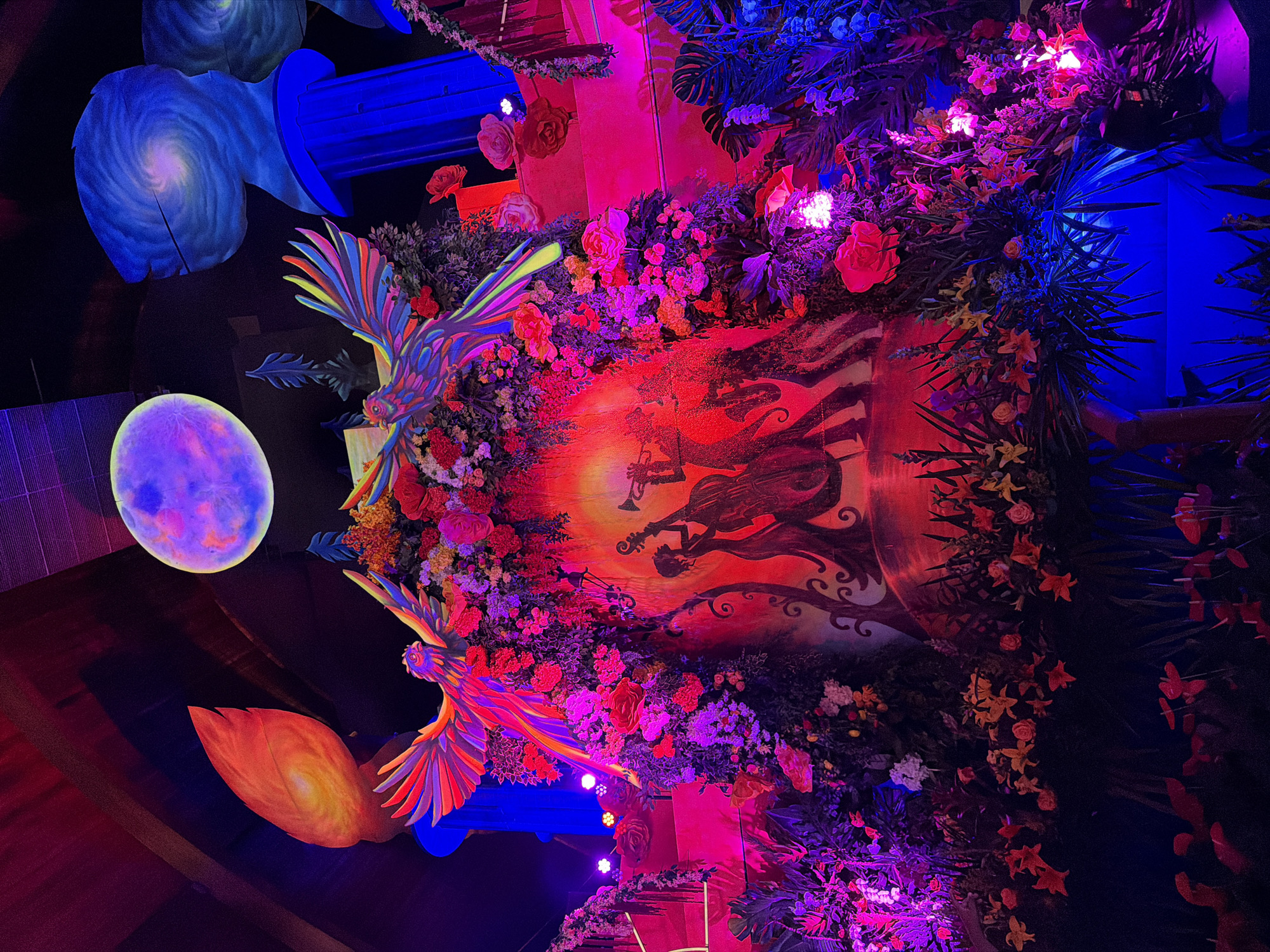The image shows a colorful and fantastical scene, likely an art installation. Two large, stylized bird sculptures with rainbow-colored wings are prominent in the foreground. They appear to be perched on or near structures resembling pillars.
Behind the birds is a backdrop featuring a large, circular, reddish-orange element with silhouettes of figures playing instruments. The overall scene is heavily decorated with artificial flowers and greenery, illuminated by various colored lights, including pink, purple, and blue.
In the periphery, there are additional decorative elements: a painted spiral galaxy and a glowing sphere that resembles a moon. The context suggests that the location might be in the city of Pomona, United States of America.