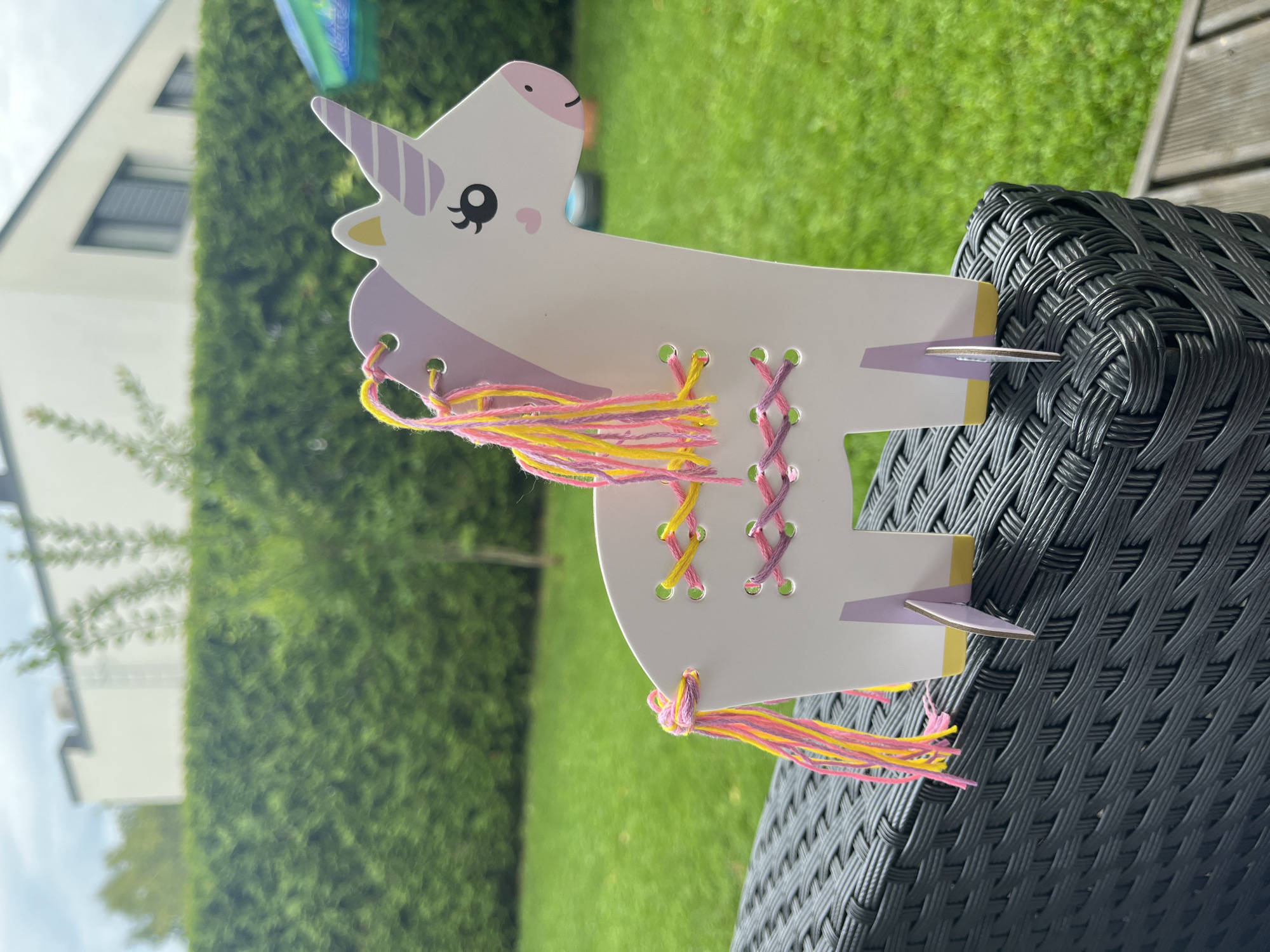 A white cardboard unicorn craft, with pink and yellow yarn threaded through its sides and tail, stands on a dark woven outdoor furniture item. The unicorn has a purple horn and mane accents, a pink heart blush, and black eyes. In the background, a manicured green hedge separates the furniture from a grassy lawn and a modern white building with dark-framed windows. The location is Haabneeme alevik, Estonia. The lighting suggests an overcast day.