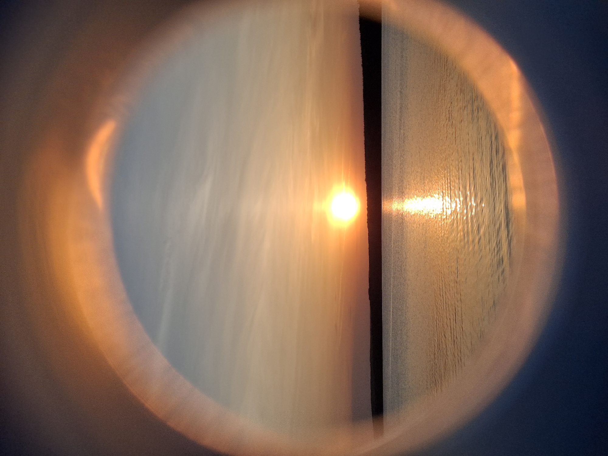 The image depicts a sunset view through a circular lens or aperture, possibly a camera lens or artistic filter. The scene shows a body of water, likely a river or lake, reflecting the light of the setting sun. The sky is visible above, with cloud formations illuminated by the golden hour light. A dark, vertical line bisects the image, possibly a structure on land or a border between two different bodies of water. The image has a blurred effect around the edges of the circular frame. The location is not directly discernible from the image content, and cannot be accurately located in Chaikovsky, Russia. No people or specific objects are identifiable.