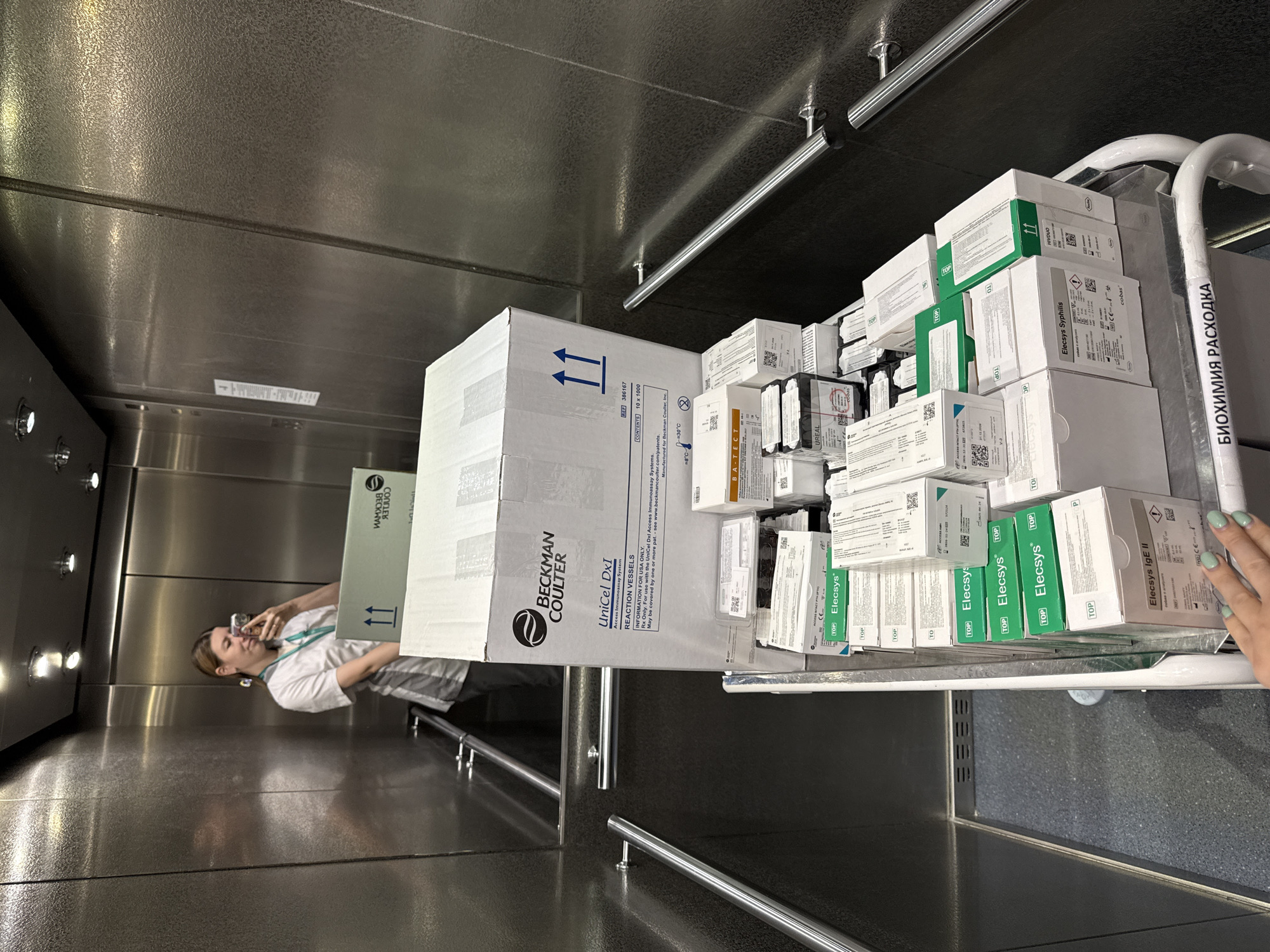 The photo shows an elevator, seemingly in Moscow, Russia, filled with boxes and a person.
Environment: The elevator's interior is metallic, likely stainless steel, with a shiny, reflective surface. There are recessed lights in the ceiling. Rails are present along the walls of the elevator.
People: A person stands inside the elevator, appearing to be taking a photo.
Objects: A large white box labeled "Beckman Coulter UniCel Dxl Access Immunoassay System REACTION VESSELS" is prominent. A cart is overloaded with smaller boxes, many of which are labeled "Elecsys" and related to various medical tests. The cart has a label in Russian, "БИОХИМИЯ РАСХОДКА", which translates to "Biochemistry Expenses" or "Biochemistry Supplies."
Actions: The person inside the elevator is taking a photo. Medical supplies are being transported, presumably for laboratory use.
Interactions: The person is likely associated with the medical supplies and their transport.