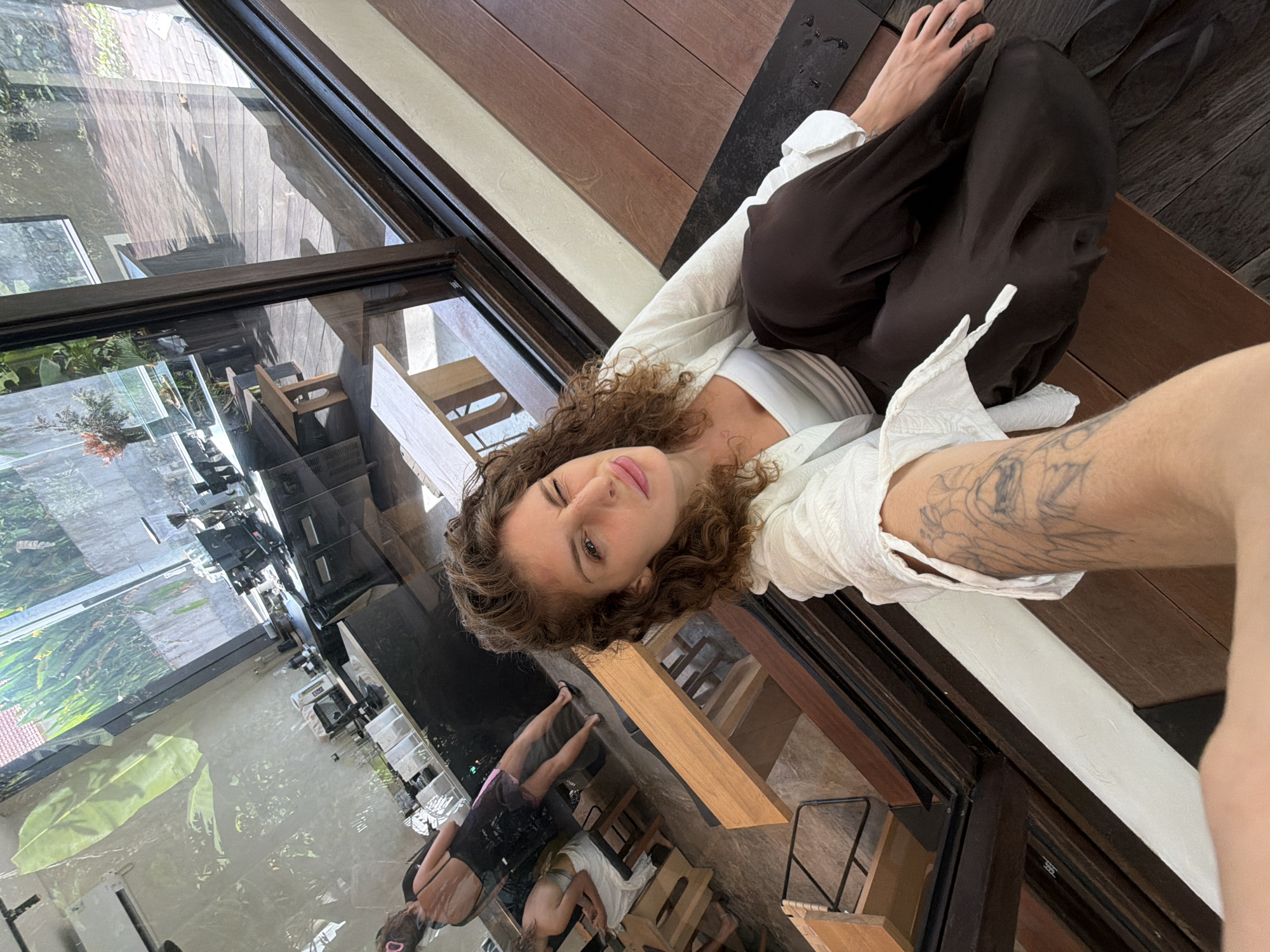 A young woman with curly brown hair is taking a selfie in what appears to be a cafe or restaurant in Mengwi, Indonesia. She is wearing a white top and dark brown pants, and a white open shirt over her top. She is sitting on a wooden bench by a large window that looks out onto a lush, green outdoor area with trees and plants. Inside the cafe, a counter with coffee machines and seating areas are visible. The woman is winking at the camera and her left arm, which is extended to take the selfie, is tattooed. Other patrons are visible in the reflection on the window, one in a black dress and another in a white top. The overall ambiance suggests a relaxed and stylish setting.