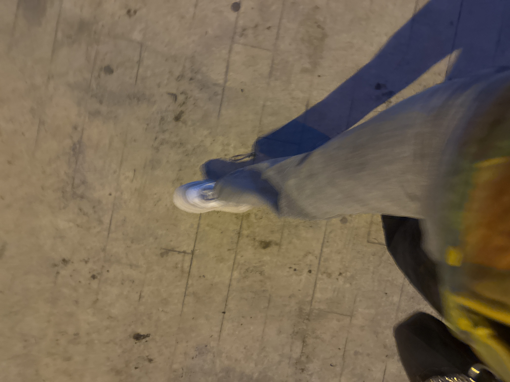 The image depicts a person's legs from a high angle, standing on a concrete surface. The person is wearing light gray jeans and white sneakers. The concrete surface appears to be outdoors, possibly a sidewalk or pavement. The lighting suggests it could be evening or night. Part of a possibly black object is visible on the right. The location is unspecified.