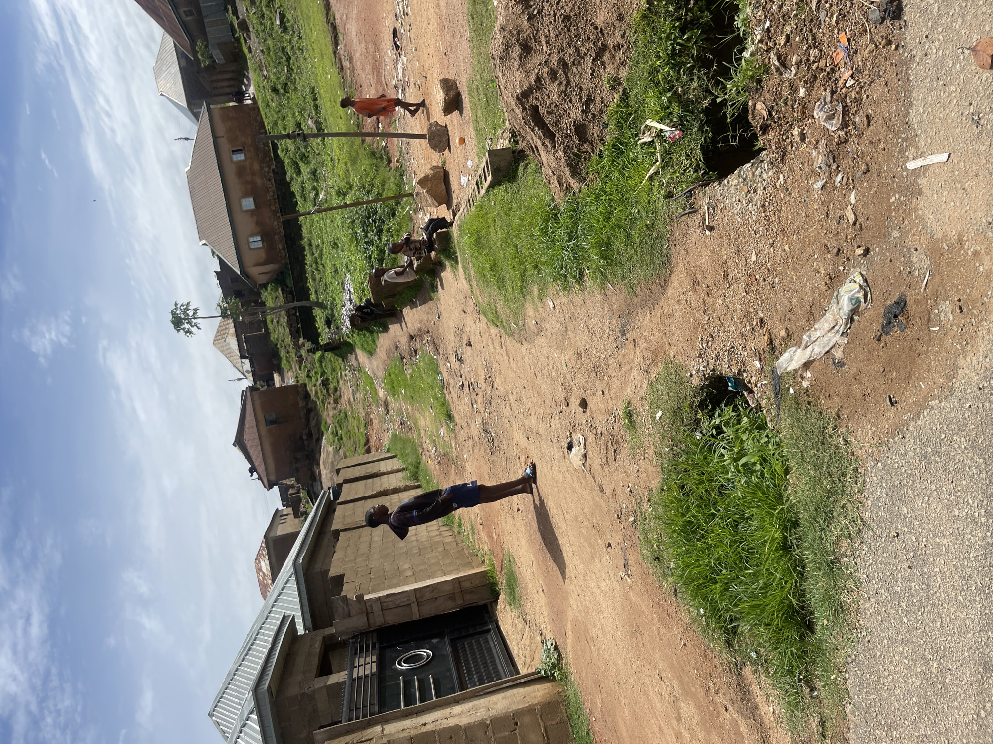 The image depicts a scene in Jos, Nigeria, on a dirt road lined with residential buildings. The buildings appear to be constructed from concrete blocks and have corrugated metal roofs, some of which are rusted. The road is unpaved and dusty, with patches of green grass growing along the sides and in a large hole in the ground. Several people are visible in the image. In the foreground, a young Black male wearing a dark hoodie and blue shorts stands facing away from the camera, leaning against a brick wall with a large metal gate. Further down the road, a group of children are gathered, some sitting on a low wall and others standing. One child, wearing an orange shirt, walks away from the camera. The environment is outdoors, under a cloudy sky. There are piles of dirt and debris scattered along the roadside.