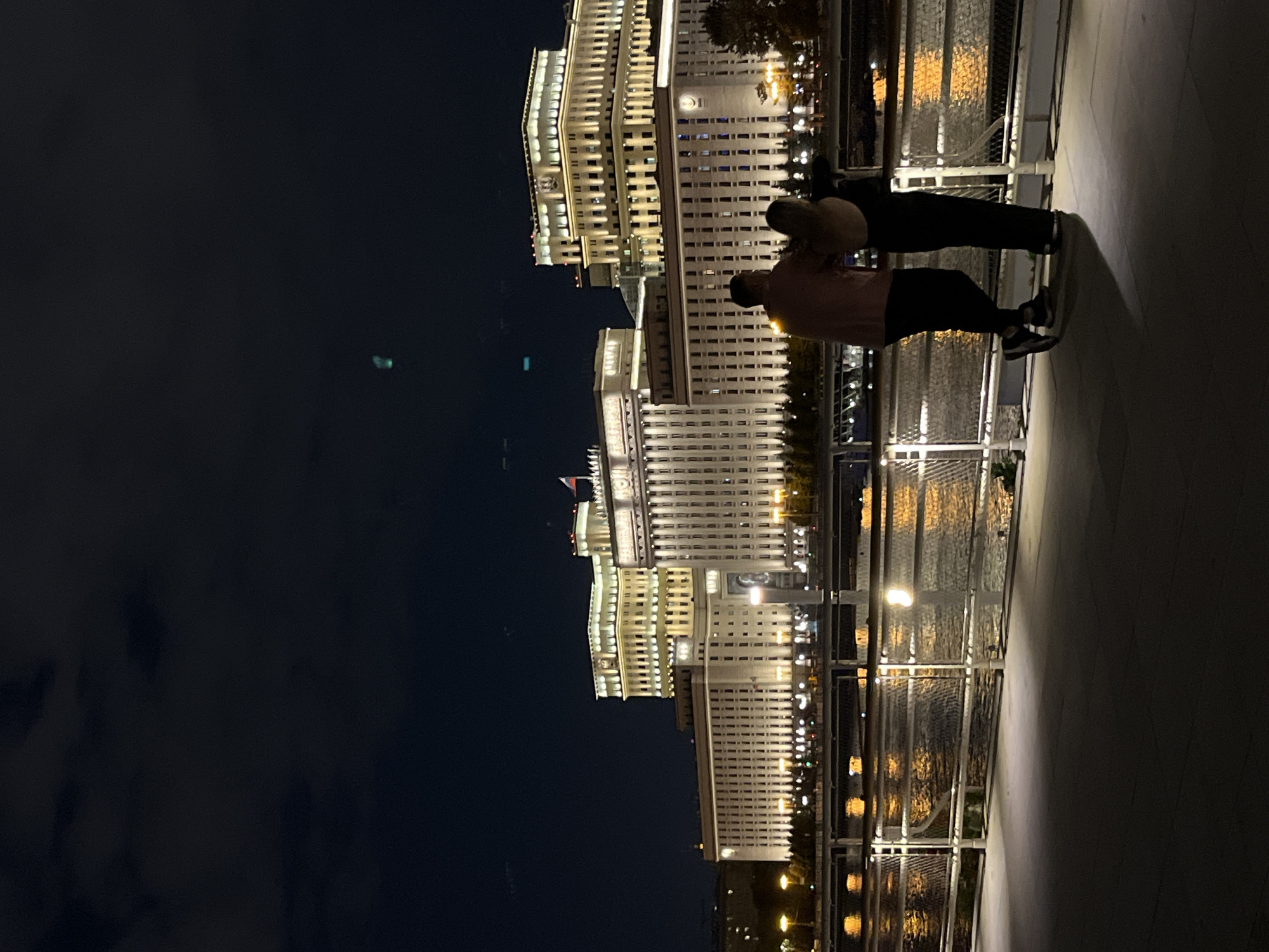 A night photograph captures a scenic view of Moscow, Russia, rotated 90 degrees counter-clockwise from its intended orientation. In the foreground, two silhouetted figures stand on a promenade or bridge, leaning against a metal railing. One person wears a light-colored top and dark pants, while the other is dressed in darker clothing. Their backs are largely to the viewer, as they gaze across a body of water towards a magnificent, illuminated building complex.
The water, likely the Moscow River or a canal, shimmers with reflections of the city lights. In the background, the grand, multi-story buildings are brightly lit, showcasing their impressive architecture, which features numerous windows and ornate detailing. One section of the complex prominently displays a clock face on a tower, while another features a flag with the distinctive white, blue, and red horizontal stripes of the Russian national flag. These architectural cues and the flag firmly place the scene in Moscow.
The sky above is dark, indicating nighttime, with subtle cloud formations visible in the deeper shadows. The overall ambiance is calm and contemplative, as the two individuals appear to be enjoying the illuminated cityscape. No discernible text is visible in the image.