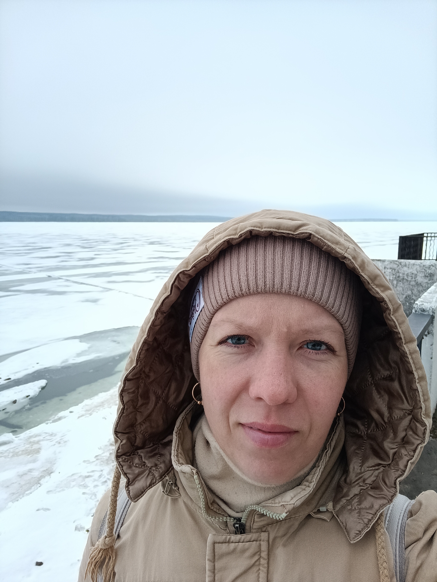 A woman is taking a selfie outdoors in Chaikovsky, Russia. She is wearing a beige beanie and a beige hooded jacket. A frozen body of water with ice floes stretches behind her to the horizon, which is obscured by an overcast sky. A short section of white concrete wall with a dark metal railing is visible to her right.
