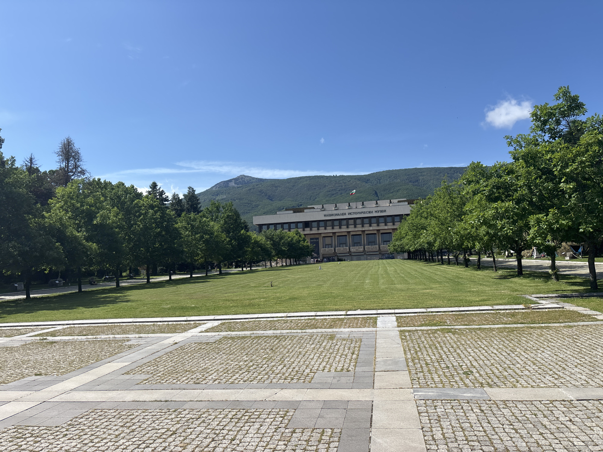 The image shows the National Historical Museum in Sofia, Bulgaria. The museum is a large, modern building with a flat roof and a facade of windows. It is surrounded by a large, well-maintained lawn and rows of trees. In the foreground, there is a paved area with a geometric pattern. The sky is clear and blue, with a few wispy clouds. The overall scene is one of order and tranquility.