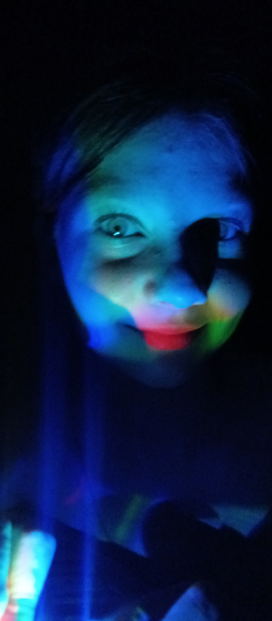 A person's face is illuminated with multicolored light, predominantly blue, red, and green, against a dark background. The location is Unknown, Unknown.