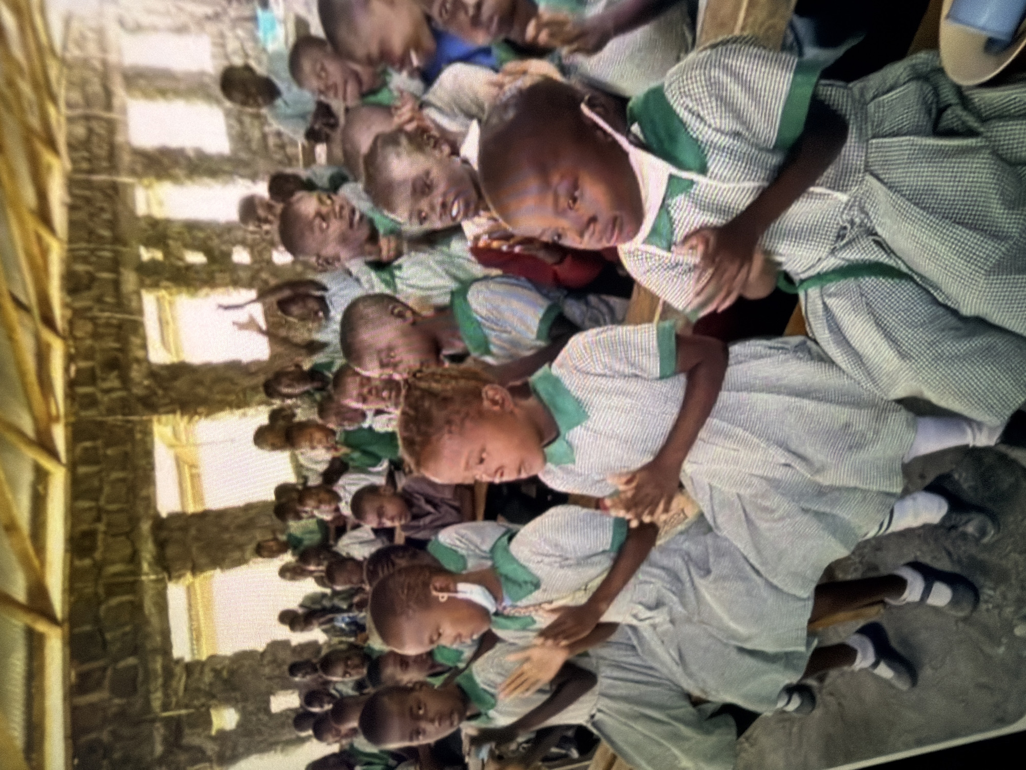 A group of children, likely students, are seated on wooden benches inside a building. The building appears to be constructed with rough, natural stone walls and a wooden roof structure. Sunlight streams in through openings in the walls, illuminating the scene. The children are wearing similar uniforms, which consist of light-colored dresses with green collars and trim, and some have masks. Many of the children are looking forward, and some have their hands clasped or raised. The overall impression is of a school or community gathering in a rural or underdeveloped setting.