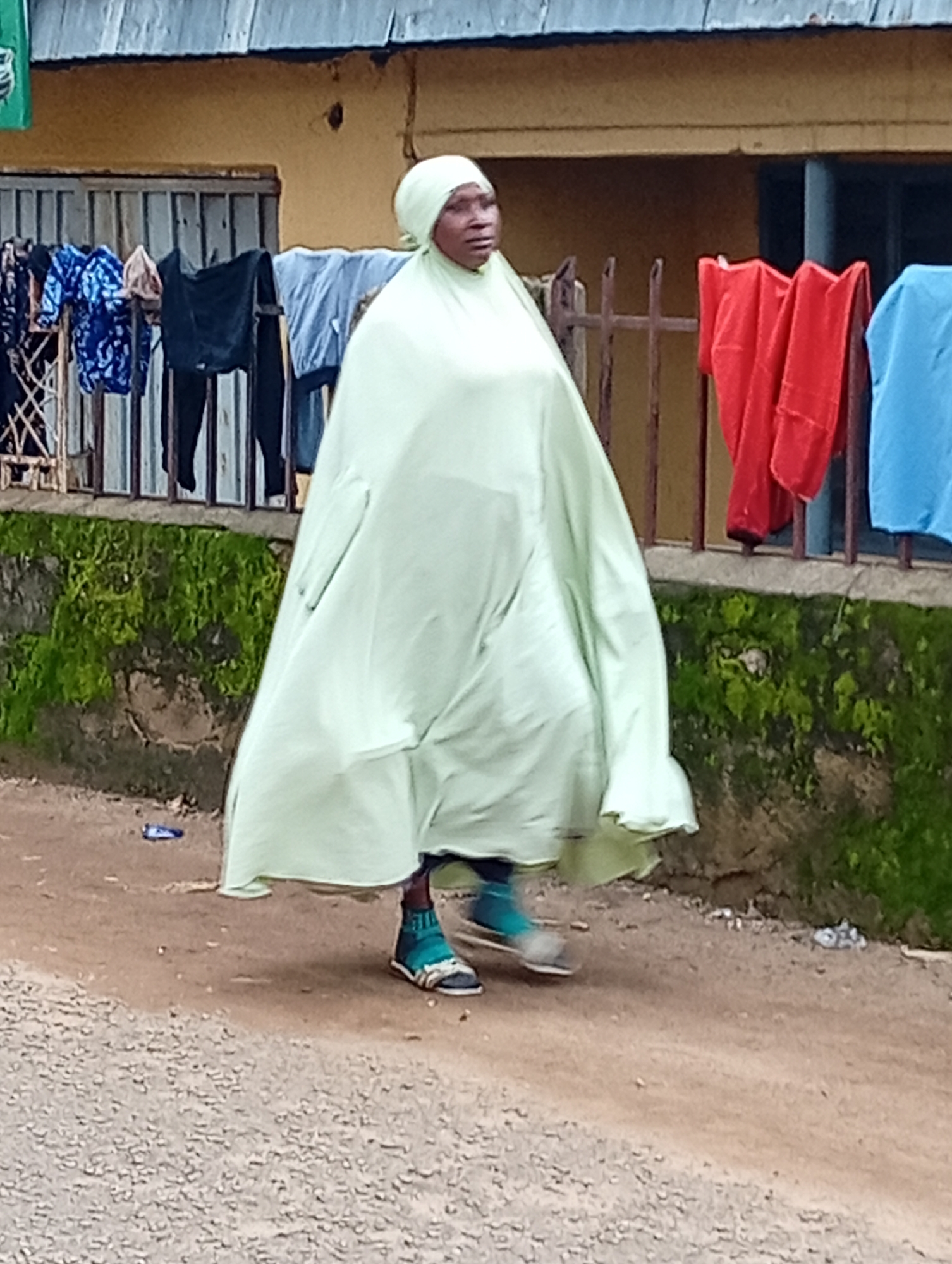 The image captures a woman walking on a dirt road in Jos, Nigeria. She is wearing a pale green hijab and a long, flowing pale green garment. Her feet are clad in teal and white sandals. Behind her, clothes are hanging on a line, and a rusty metal fence separates the road from a building with a yellow facade. Green moss covers a stone wall to the left of the fence. The ground is a mix of dirt and gravel, with some scattered debris, including a blue plastic object. The sky is overcast.