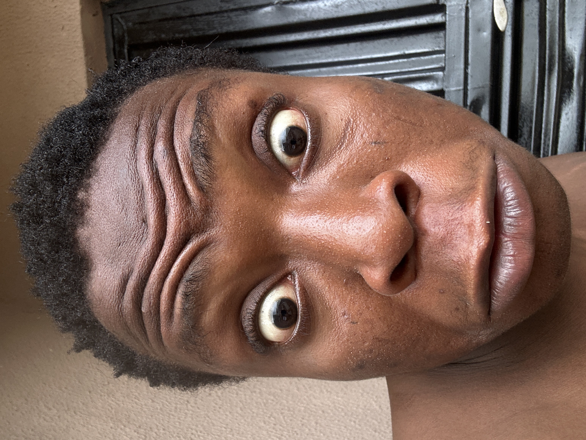 The image is a close-up, tilted portrait of a Black male. The subject's face is in sharp focus, revealing details of his skin texture, wrinkles on his forehead, and the irises of his eyes. He is looking directly at the camera with a somewhat surprised expression. His hair is short and curly. Part of his bare chest is visible at the bottom of the frame. The background is slightly blurred, showing a wall and a dark metal structure with horizontal lines. The location in the city is unknown.