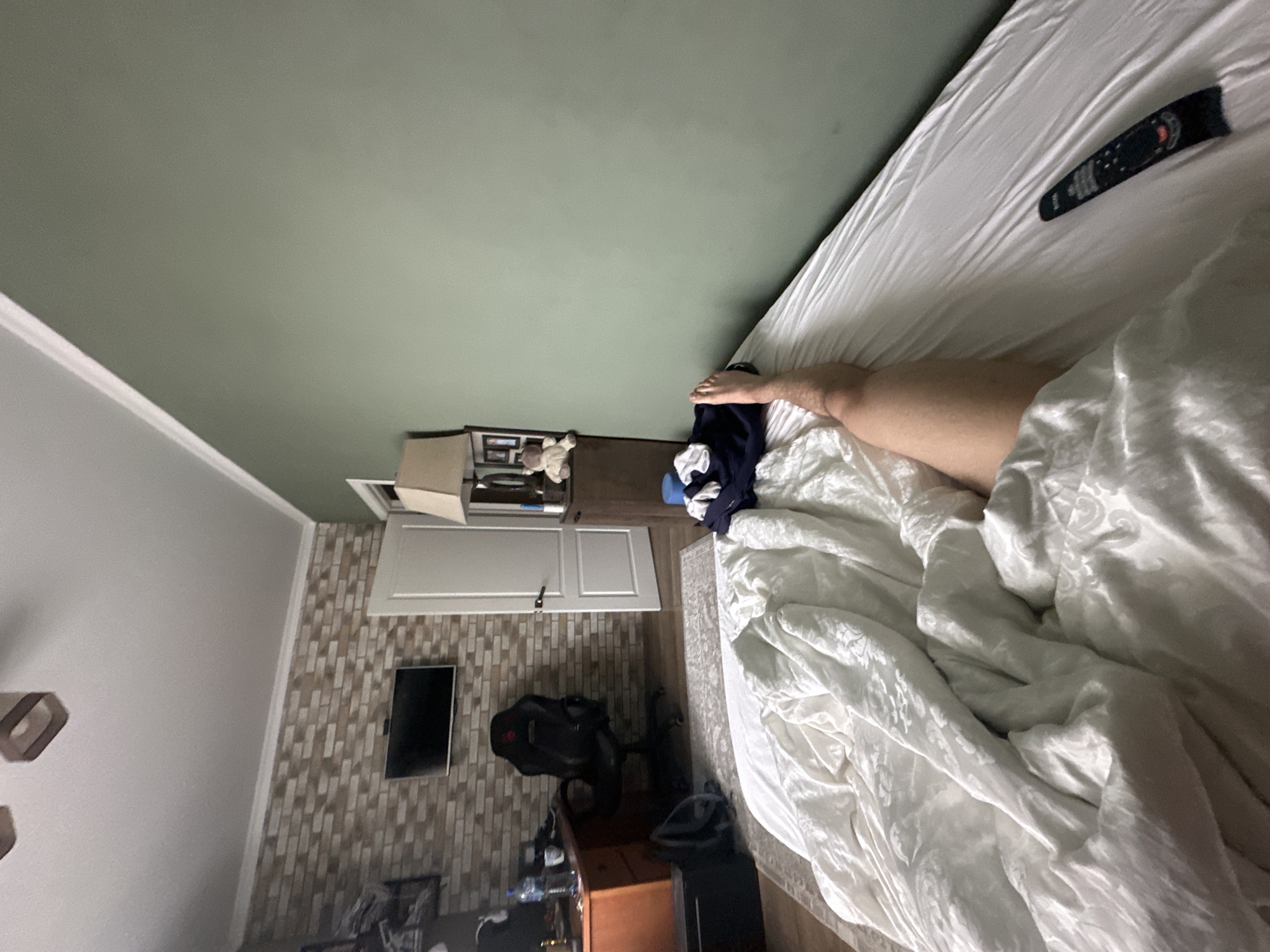 The image depicts a bedroom interior, captured from a low angle looking upwards and across the room. A bed, covered in rumpled white bedding with a subtle patterned texture, occupies the right side of the frame. A bare leg and foot extend from beneath the covers, suggesting a person is resting or just waking up. A black remote control with red and white buttons lies on the bed near the foot area.
The floor, visible on the left and center, is dark wood or laminate, partly covered by a light-colored rug next to the bed. A pile of dark clothing is on the floor near the bed. The wall on the left features a textured brick-like pattern, above which is a plain white wall with white crown molding. A flat-screen television is mounted on the brick-patterned wall, and a white door, slightly ajar, is also seen there.
In the lower left, a wooden desk or dresser holds a water bottle and other items, with a black gaming or office chair featuring red accents positioned in front of it. A small nightstand or dresser is situated next to the bed, displaying framed pictures, a plush toy, and a box. The ceiling, painted a light sage green, dominates the upper portion of the image. The scene suggests a relaxed atmosphere in a home setting during what appears to be daytime. No clearly legible text is visible on any objects.