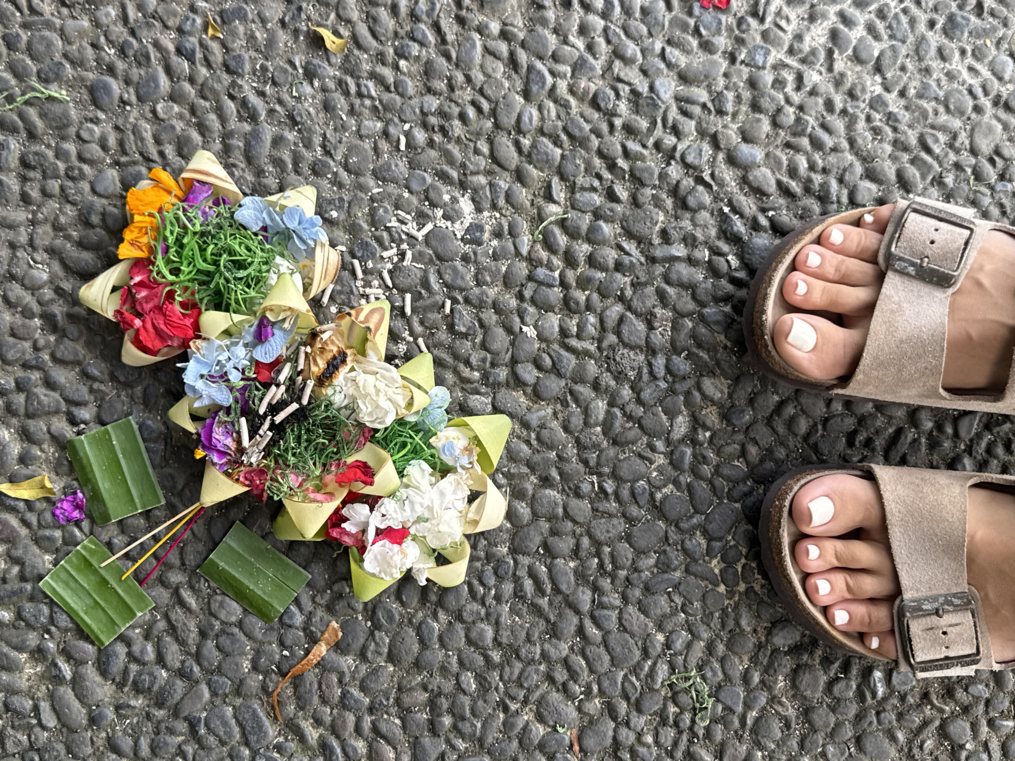 A high-angle medium shot shows a section of a grey cobblestone walkway in Kuta Selatan, Indonesia. A colorful Balinese offering, a Canang sari, sits on the left side of the frame. The offering is made of woven coconut leaves and filled with flowers, incense sticks, and other symbolic items. Scattered around the offering are bits of rice and flower petals. On the right side of the frame are the feet of a person wearing beige Birkenstock-style sandals. The person's toenails are painted white. The cobblestone walkway provides a textured background.