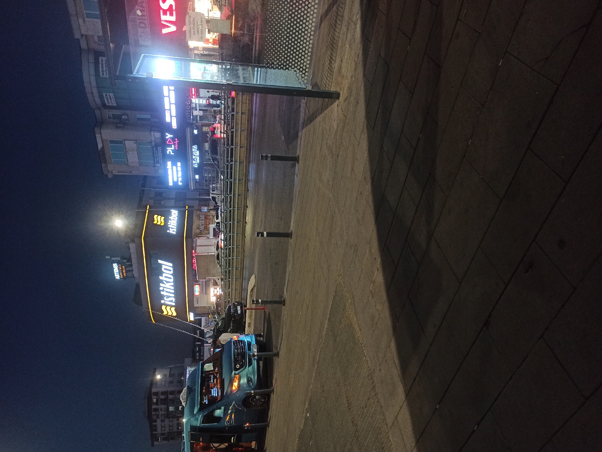 This image captures a nighttime street scene in Ümraniye, Turkey, specifically near the "İstikbal" furniture store. A light blue minivan with its door open is parked on the sidewalk. Several bollards are placed along the edge of the sidewalk and on the street. In the background, a multi-story building features illuminated signs for "İstikbal," "ARTEMIS Düğün Halo Salonları" (Artemis Wedding Hall), and other businesses. Across the street, there are more illuminated signs, including "VES" and "DICA." The street is paved with asphalt and appears to be a busy road with several vehicles, including a black car. The overall lighting suggests it is nighttime, with artificial lights from the businesses and streetlights illuminating the scene.