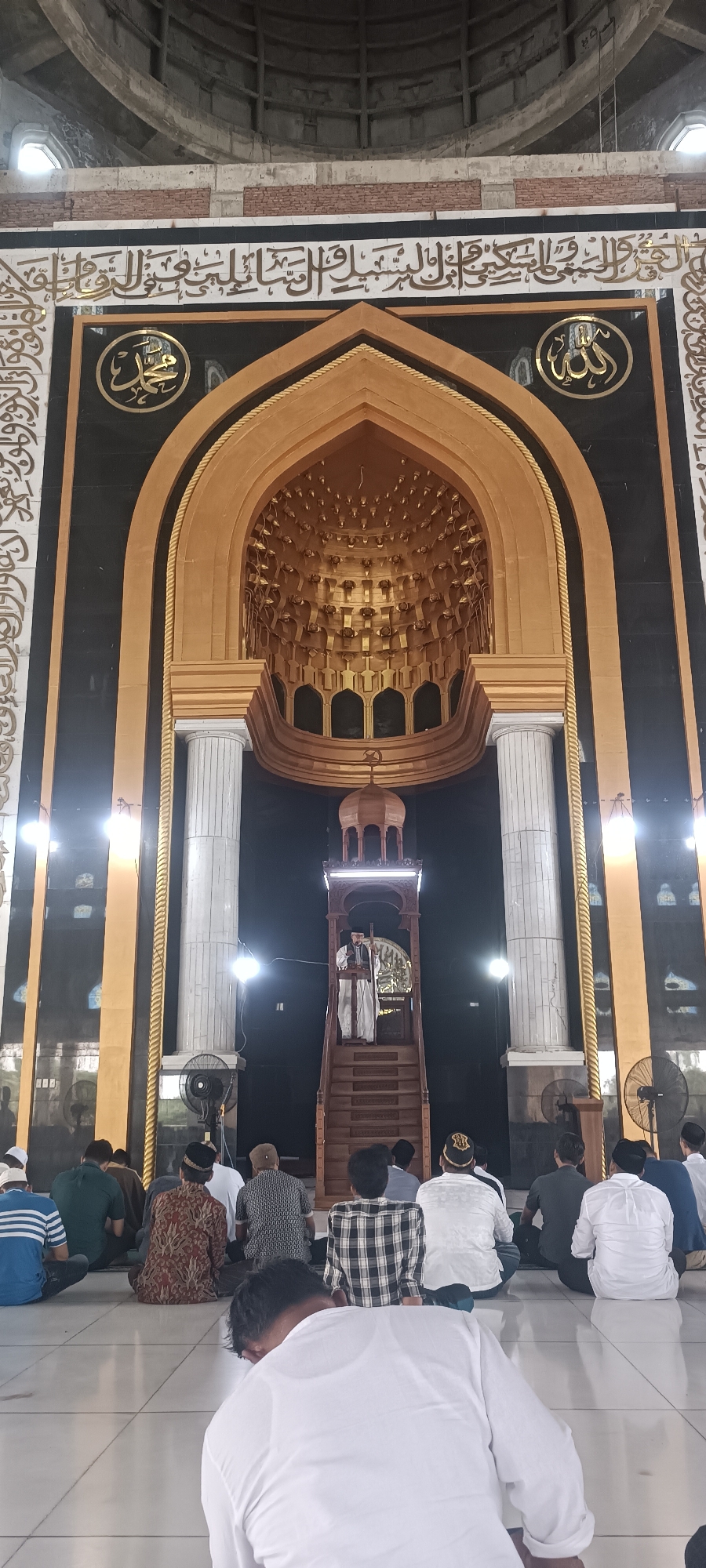 The image captures an interior view of the Baiturrahman Grand Mosque in Lhokseumawe, Indonesia, during a prayer service. A man in traditional Islamic attire stands on a wooden pulpit, addressing the congregation. He is wearing a white robe and a black turban. The pulpit is intricately carved and elevated, with a wooden dome structure above it.
In front of the pulpit, a group of men are seated on the tiled floor, facing the pulpit. They are dressed in various attire, including sarongs, shirts, and caps, indicating a communal prayer. The mosque's interior features grand architecture, with large white columns flanking the pulpit. The walls are adorned with Arabic calligraphy and intricate golden patterns on a dark background. The ceiling appears to be a dome with a complex geometric design in gold. Lighting is provided by overhead fixtures, casting a warm glow on the scene. The overall atmosphere suggests a solemn and respectful religious gathering.