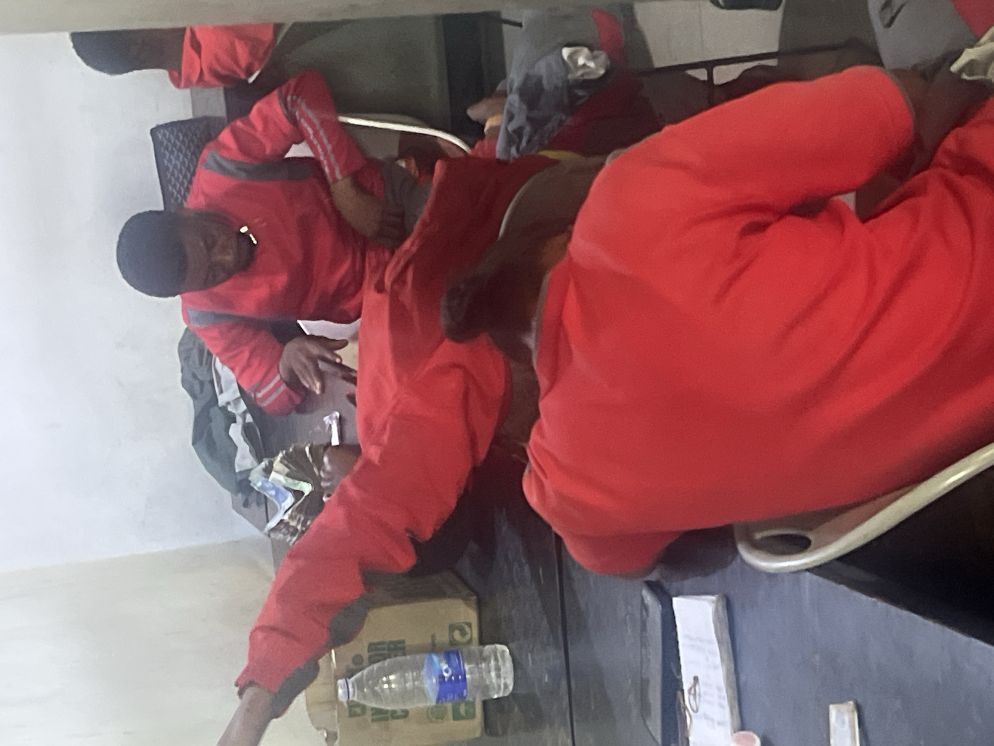 The image displays an indoor scene featuring several individuals, predominantly dressed in red tracksuits or similar casual wear, in what appears to be a common room or waiting area. The photograph is rotated 90 degrees clockwise.
In the foreground, a person in a full red tracksuit is seen lying down across two chairs or a bench, their body extending from the left to the right side of the frame. Another individual, also wearing a red tracksuit with gray stripes on the sleeves, is seated at a dark table or desk on the left, looking downwards. Their hands are resting on the table, near what appears to be a small pile of currency notes, likely Nigerian Naira given the context. Other people in red clothing are partially visible in the background, suggesting a group gathering. One person's legs are visible under a table to the right, also in red attire.
Notable objects on the table surfaces include a plastic water bottle with the brand "AQUA" clearly visible on its label, placed on a cardboard box, and some papers or notebooks. The walls of the room are light-colored, indicating an indoor environment. The uniform-like red clothing suggests that the individuals might belong to a specific group, team, or institution. The overall atmosphere appears to be one of resting or waiting. There are no direct cues for the time of day other than artificial indoor lighting.