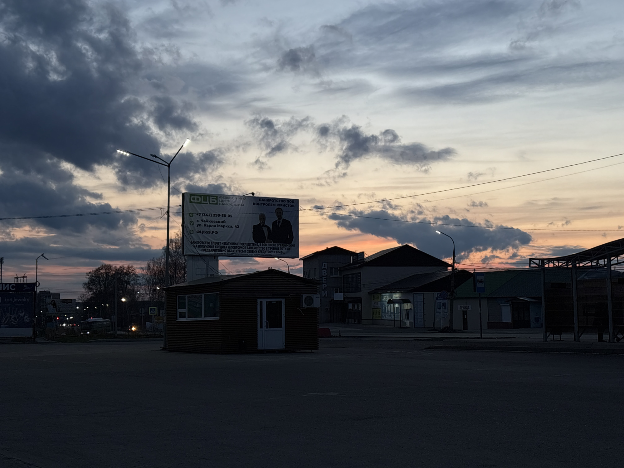 The image captures a street scene in Chaikovsky, Russia, at dusk or dawn. The sky is filled with dramatic clouds tinged with the colors of the sunrise or sunset.
Dominating the foreground is an empty asphalt lot, partially obscured by shadow. In the middle ground, there's a small, rectangular wooden building, possibly a kiosk or guard station. Behind it, a large billboard advertises "ФЦБ", with text in Russian indicating bankruptcy services and contact information including the phone number +7 (342) 299-50-05 and the address "г. Чайковский ул. Карла Маркса, 42". The billboard also features images of two men in suits.
Further back, various buildings line the street, including shops with visible signage in Russian. Streetlights are illuminated, adding to the ambiance. The buildings appear to be of standard urban construction for a Russian city. A bus stop is visible on the right side of the image.
The overall impression is one of a quiet, somewhat desolate urban landscape. The time of day suggests either early morning or late evening, contributing to the stillness of the scene.