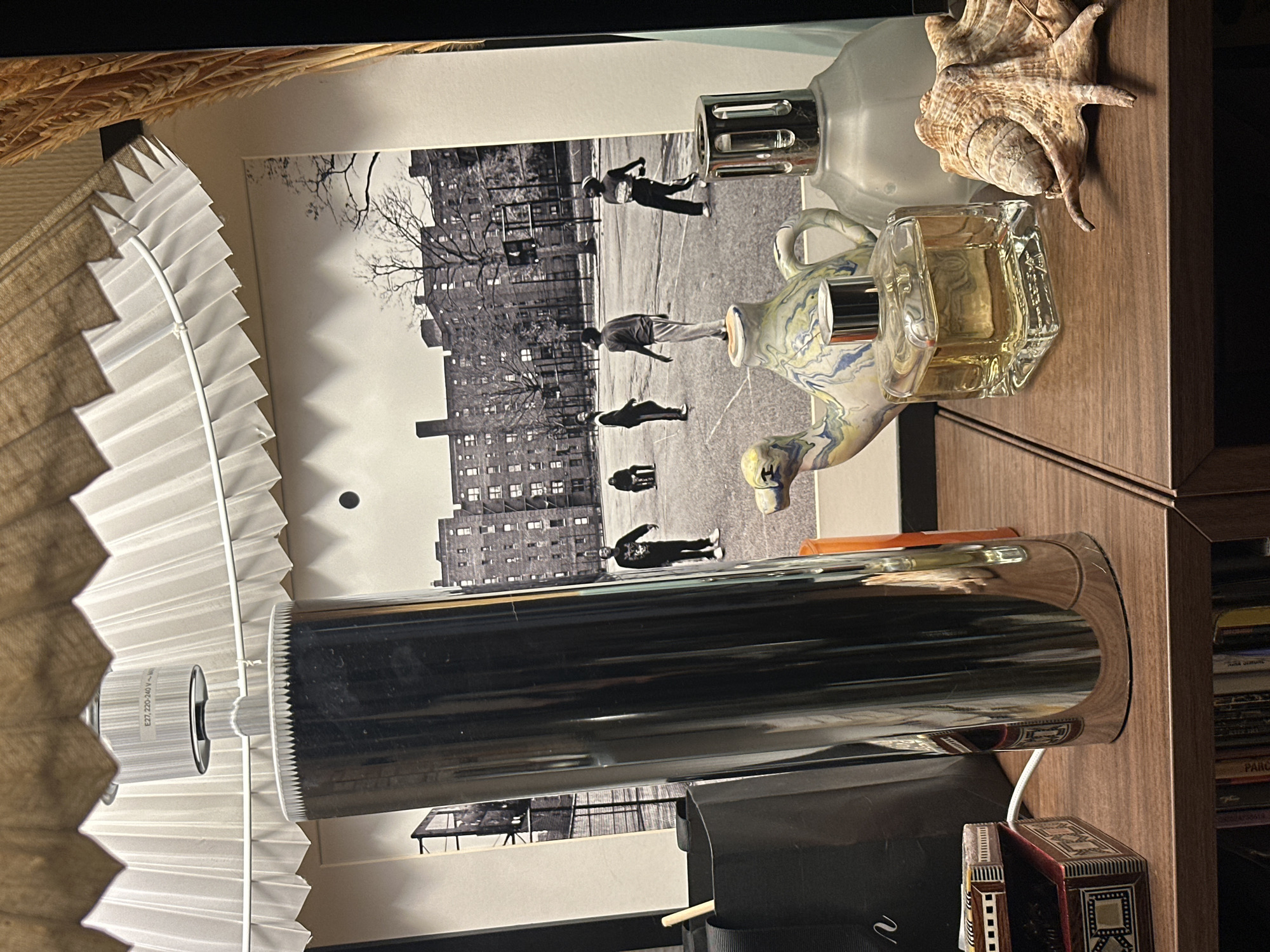 The scene depicts an indoor tabletop or shelf display, located in a residence in Paris, France.
In the foreground, on a dark brown wooden surface, multiple objects are arranged. To the left, a small, open, ornate wooden box with intricate inlay patterns holds a harmonica. A white cable is visible near its base. Behind this box, a black bag with a partially visible white logo and a light-colored stick inside is present. Dominating the center-left is a tall, cylindrical, reflective silver-metallic object, likely a lamp base, reflecting parts of the room and the background image. An orange cylindrical votive or candle holder is partially obscured behind it.
To the right, a collection of decorative and functional items is arranged. These include a ceramic teapot-like object featuring a blue, white, and yellow marbled pattern, with a silver cap resting on its spout. Next to it, a square glass bottle containing amber-colored liquid, sealed with a silver cap, is visible, likely a perfume bottle. Further right is a frosted glass bottle with a decorative metallic silver top, characteristic of a catalytic lamp (e.g., Lampe Berger). On the far right, a large, intricate, beige and brown conch seashell with a textured surface rests on the wooden surface.
In the background, a large, framed black and white photograph is prominently displayed. This photograph depicts an outdoor urban basketball court scene. Five individuals, appearing as young males in casual clothing, are scattered across the court; four are standing or in motion, while one is seated on the far left. A dark circular object, a basketball, is visible in mid-air above the court. The background of the photograph shows multi-story residential buildings, likely brick or stone, characteristic of an urban housing complex, with numerous windows and bare trees, and a chain-link fence separating the court from the buildings.
Above the entire display, a pleated lampshade is visible, featuring an inner white pleated layer and an outer brownish, textured layer. The lamp's socket, labeled "E27, 220-240 V ~", is visible at the top.