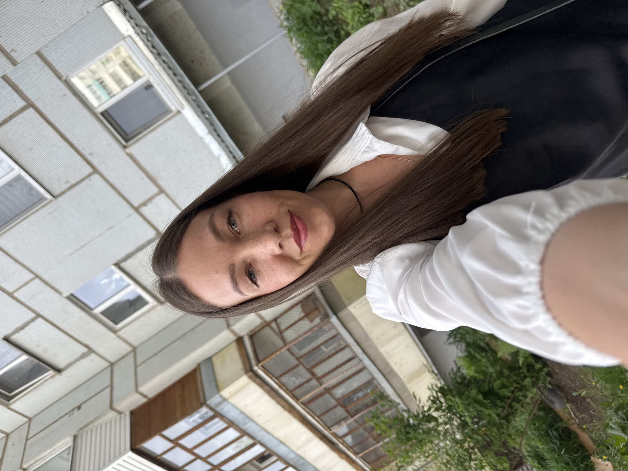 A selfie image showing a young woman with long brown hair, light skin, and freckles on her face. She is wearing a white shirt and a black vest. Her location is in Chaikovsky, Russia, as indicated by the background architecture, which includes a Soviet-era apartment building with grid-like concrete panels and small, square windows. There are some trees and vegetation visible in the foreground and beside the building. The image is taken from a slightly low angle, with the woman holding the camera up and towards herself.
