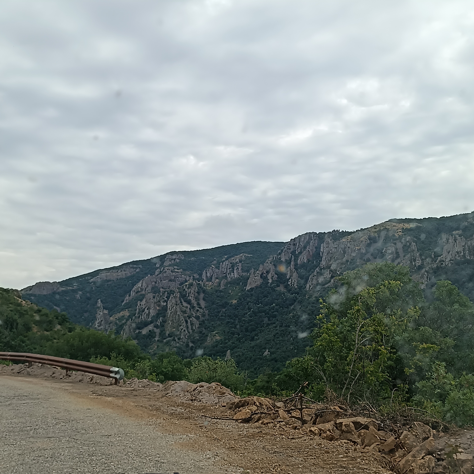 A rural road winds through a mountainous landscape under an overcast sky. The road surface is gravelly, with a rusted guardrail bordering the left side. Beyond the road, the terrain slopes upward, covered in dense green vegetation and rugged rock formations that characterize the mountains. The sky is filled with heavy, grey clouds, suggesting an impending or recent rain. The scene appears to be captured from a vehicle traveling along the road, offering a view of the natural environment of the region surrounding Madzharovo, Bulgaria.