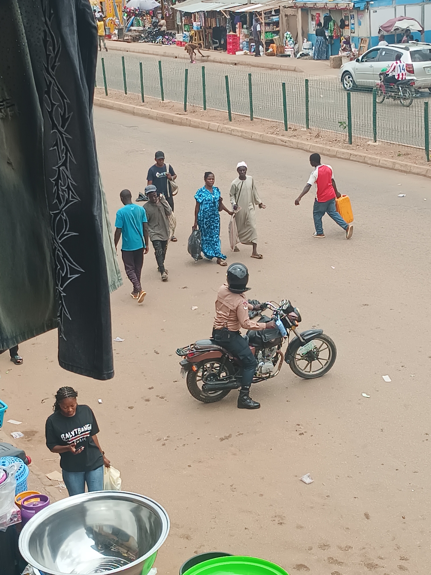 The image depicts a street scene in Bukuru, Nigeria. Several people are walking on the street, including a man in a red shirt carrying a yellow container and a woman in a blue dress. A person in a tan uniform is riding a motorcycle. A woman wearing a black t-shirt and jeans is looking at her phone while standing near a collection of plastic buckets and a metal bowl. A silver car is parked on the side of the road, and a fence separates the street from a market area with stalls and people. The street surface is dusty and light brown.