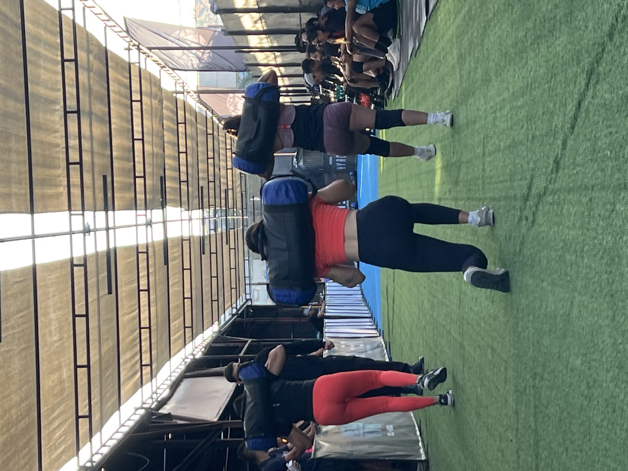 Three individuals are engaged in a fitness activity, carrying large weighted bags across their shoulders on a green turf surface. The scene appears to be an outdoor gym or training facility, possibly located in the city of Arica, Chile, indicated by the architecture and the open-air setup with a covered roof supported by metal beams.
The person in the foreground, viewed from behind, is wearing a bright orange cropped top and black leggings, with a weighted bag on their shoulders. Their right knee is bent as if in mid-lunge or stepping motion. To their left, another individual in a black top and bright red leggings is also carrying a similar weighted bag. A third individual, slightly further back and to the right, wears a dark top and burgundy shorts with knee pads, also carrying a weighted bag.
In the background, several other people are seated, observing the activity. The environment is characterized by a green artificial turf floor, a covered overhead structure made of translucent material stretched over a metal framework, and various gym equipment or barriers along the sides. The lighting suggests it is daytime, with sunlight filtering through the covered roof, creating shadows and highlights.