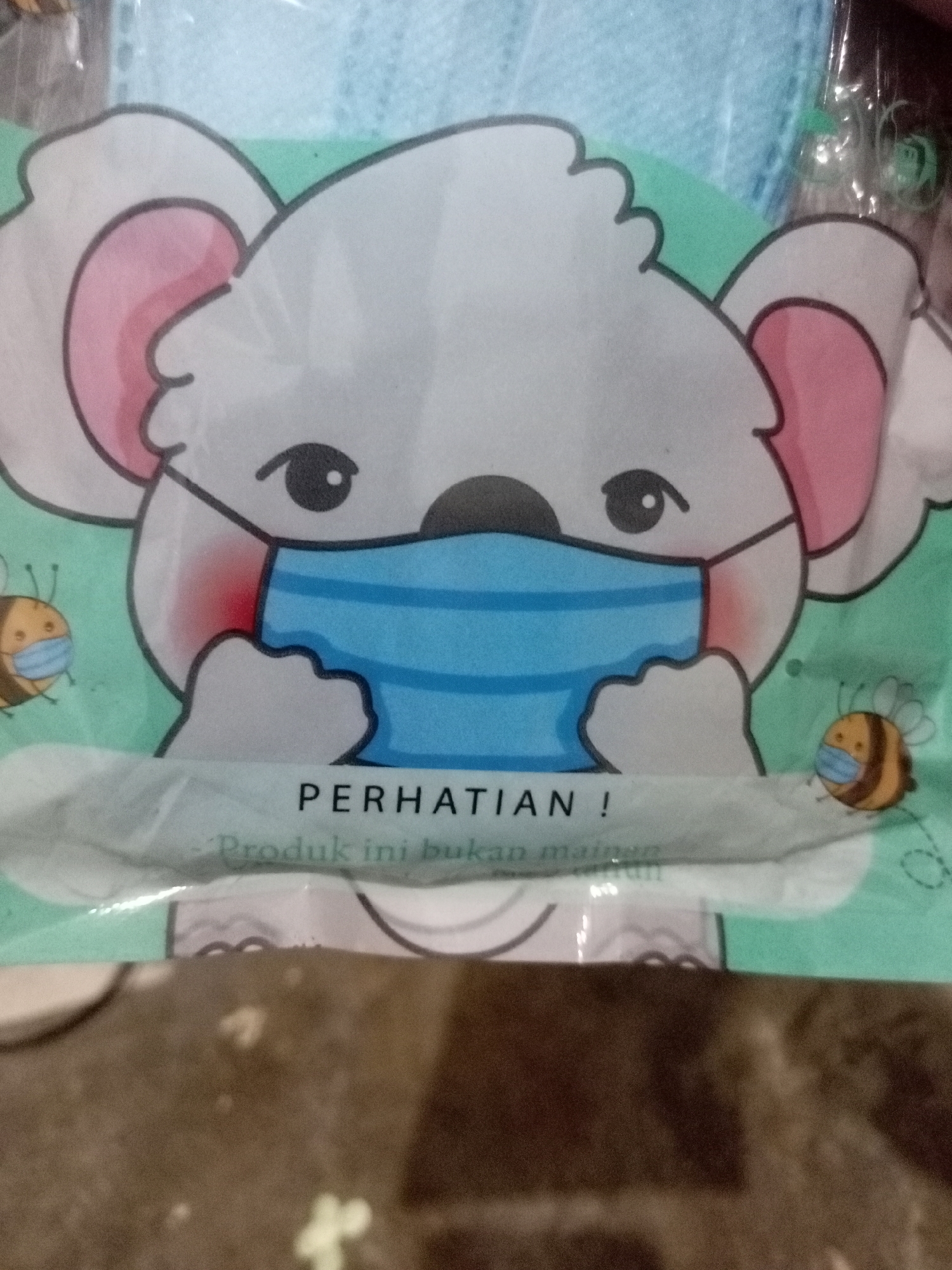 An image shows a close-up of a blue surgical mask package featuring a cartoon illustration of a white koala wearing a blue mask. Two small bees, also wearing masks, are depicted on either side of the koala. The text "PERHATIAN !" (Attention!) is prominently displayed above smaller text that reads "Produk ini bukan mainan" (This product is not a toy). The background of the packaging is a light green color. The image does not provide any specific information about its location in Batam, Indonesia.