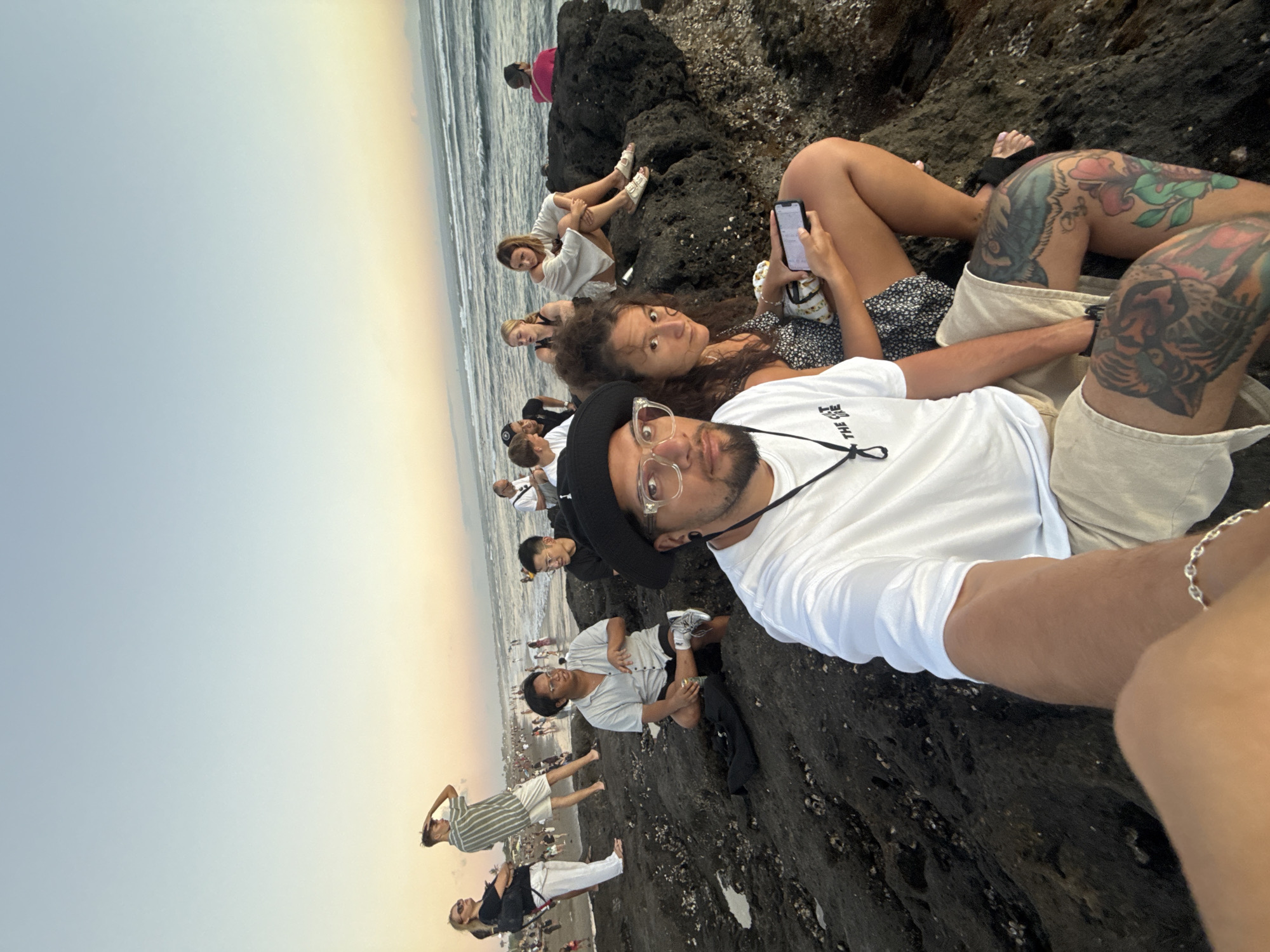 A vibrant selfie captures a group of people relaxing on a dark, rugged, volcanic-like rocky beach during the golden hour, indicative of either sunrise or sunset. The sky displays a soft gradient, transitioning from light blue at the top to warm yellow and orange hues near the horizon, which meets the ocean with gentle waves.
In the foreground, a man wearing a white t-shirt with "THE CURE" text, clear glasses, and a black hat or beanie, takes the photo. His arm, adorned with a silver chain bracelet, is partially visible. Beside him, a woman with curly hair looks towards the camera, while another person's legs, featuring distinctive and colorful floral and tiger tattoos, are prominent. This individual holds a smartphone, likely engaging with its screen.
Several other individuals are scattered across the dark rocks in the midground, some seated and others standing, enjoying the coastal view. A woman in a white outfit and another in a black top are visible among them. In the background, a larger crowd of people is seen along the sandy shoreline and closer to the water's edge, stretching along the beach. The overall atmosphere appears relaxed and social, as people gather to appreciate the serene coastal environment, typical of North Kuta, Indonesia.