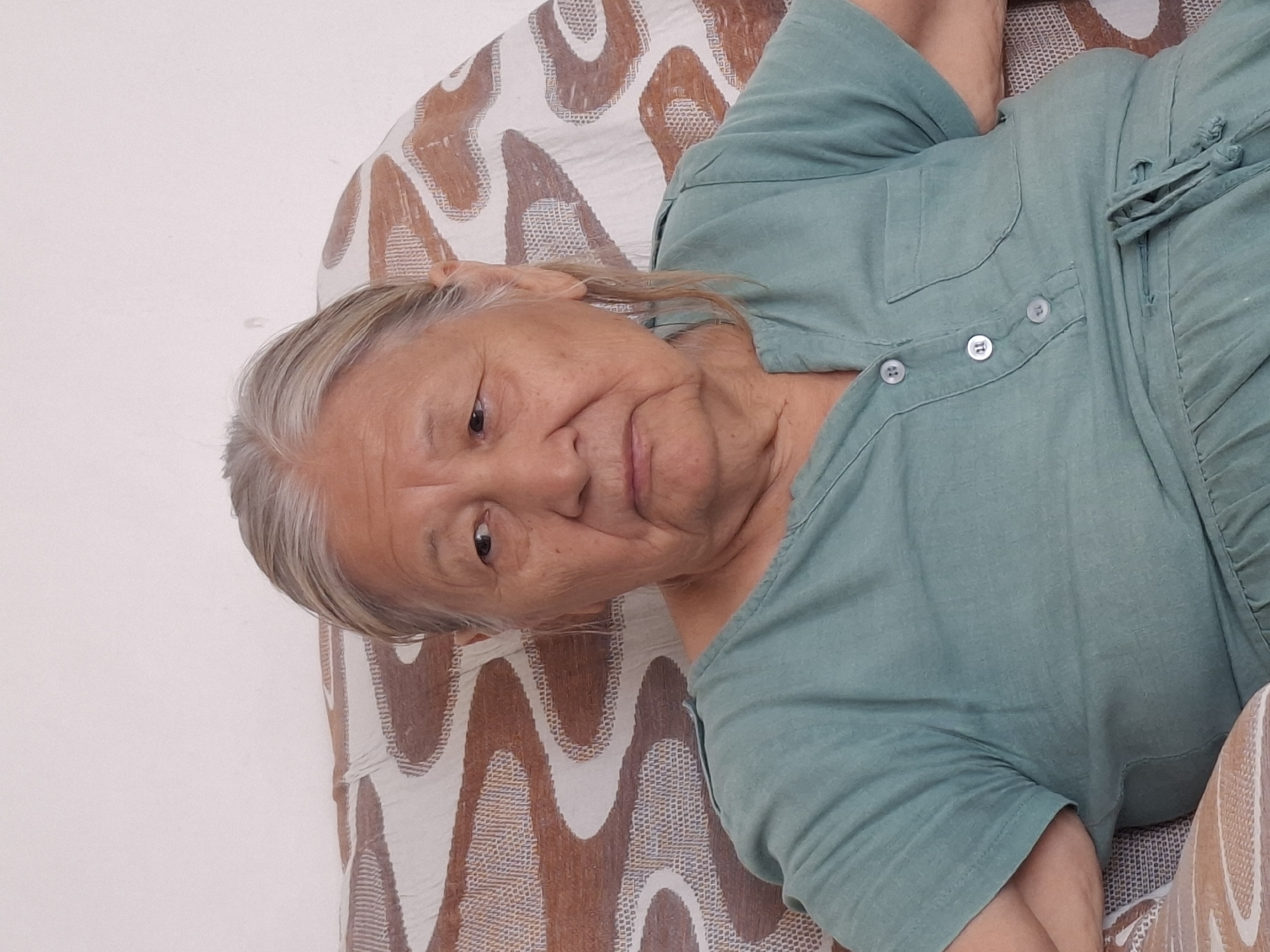 An elderly East Asian woman with grey hair is seated on a patterned sofa, looking to her right. She is wearing a teal colored button-down shirt dress. The woman is positioned in the center of the frame, with the sofa and a white wall behind her. The image appears to have been taken indoors, possibly in a residential setting. There is no indication of the specific location within the city of Almaty, Kazakhstan.