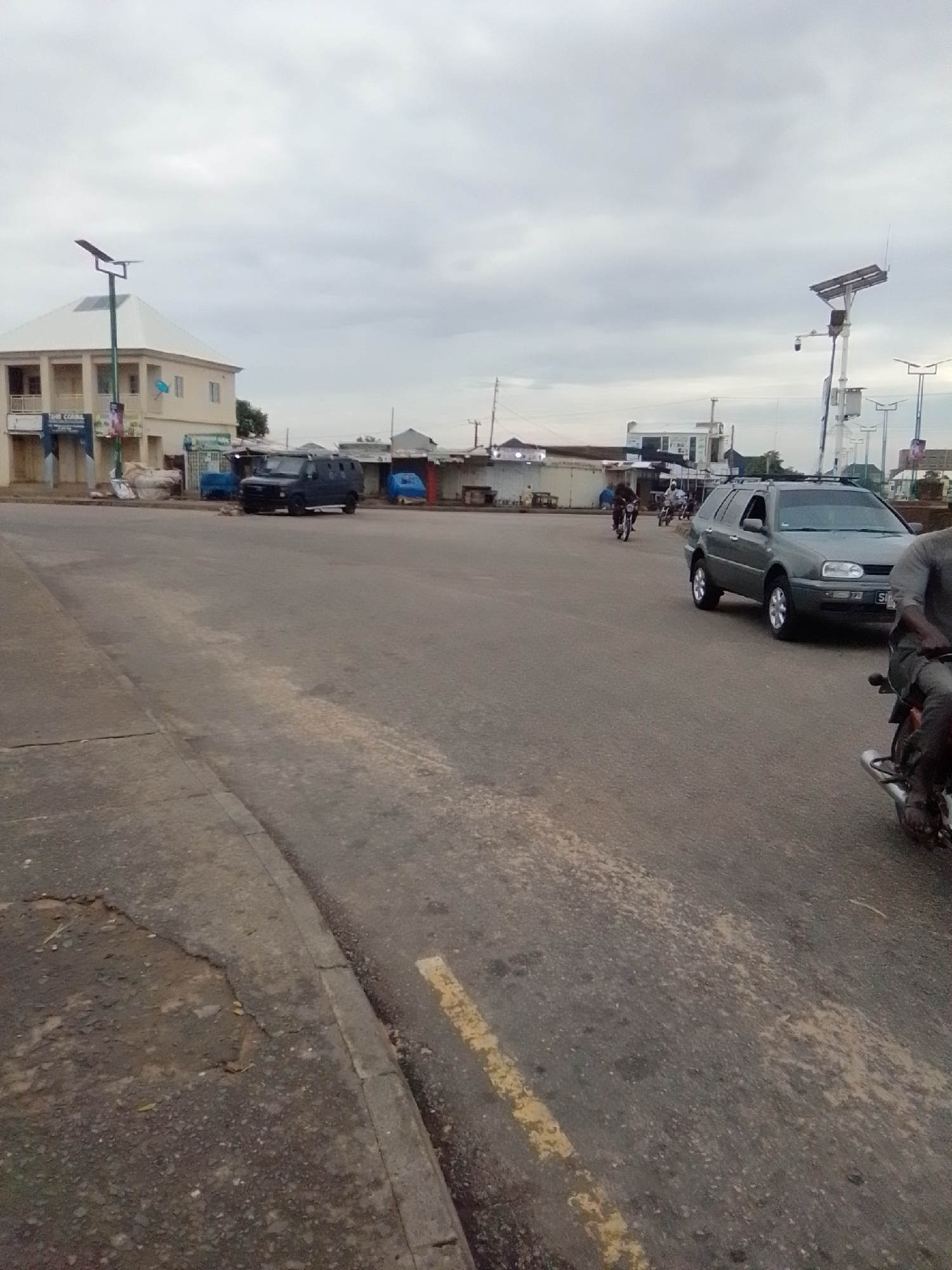 The image depicts a street scene in Pantame, Nigeria. The sky is overcast with clouds. On the left side of the road, there is a light-colored building with a white roof and a solar-powered street lamp in front of it. Adjacent to this building are several market stalls with corrugated metal roofs, some covered with blue tarps. A dark-colored armored van is parked on the side of the road in front of one of these stalls.
In the center of the image, a grey Volkswagen Golf station wagon is driving on the road. To the right of the station wagon, a person on a motorcycle is visible, with only their legs and the motorcycle's rear visible. Further in the distance, other motorcycles can be seen on the road, and more market stalls line the other side of the street. The road surface appears to be asphalt with visible patches and a faded yellow lane marking. There are also several other solar-powered street lamps visible in the background.