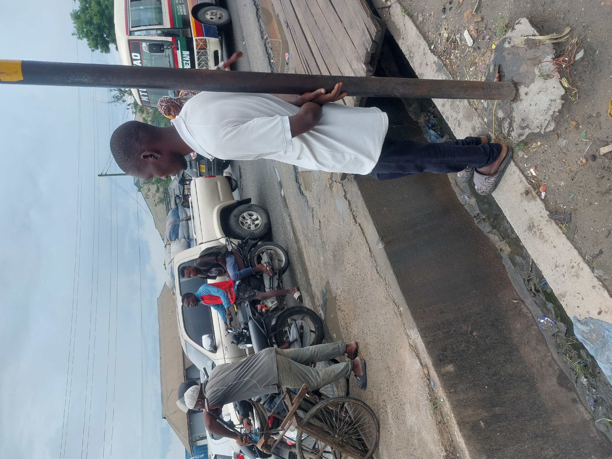 The image captures a street scene in Dar es-Salaam, Tanzania. A man in a white shirt and dark pants is standing beside a metal pole, positioned near a concrete drainage ditch filled with debris. Several individuals are visible: one working on a bicycle, another on a motorcycle, and others near a van. A bus with "REZANI" and "MTO AO" text is partially visible in the background. Overheard are the powerlines.