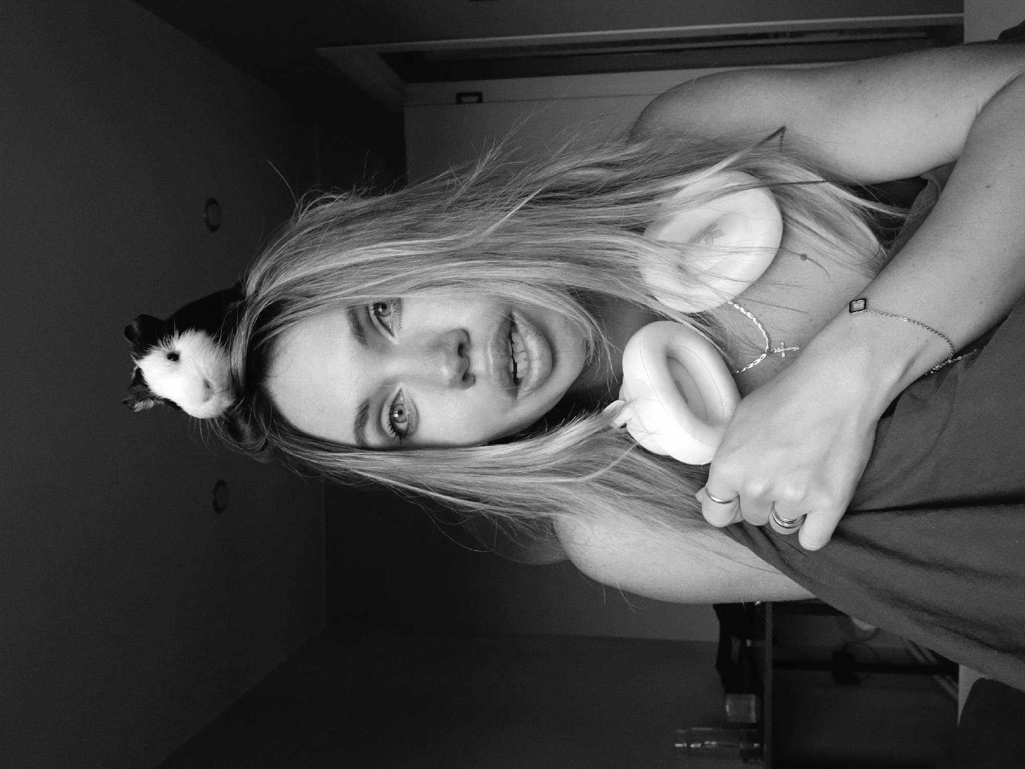 A black and white portrait shows a woman with blonde hair looking at the camera, holding white headphones. A black and white guinea pig sits on her head. A necklace and bracelet are visible on her neck and wrist. The location in the city of Votkinsk, Russia, cannot be determined from the image.