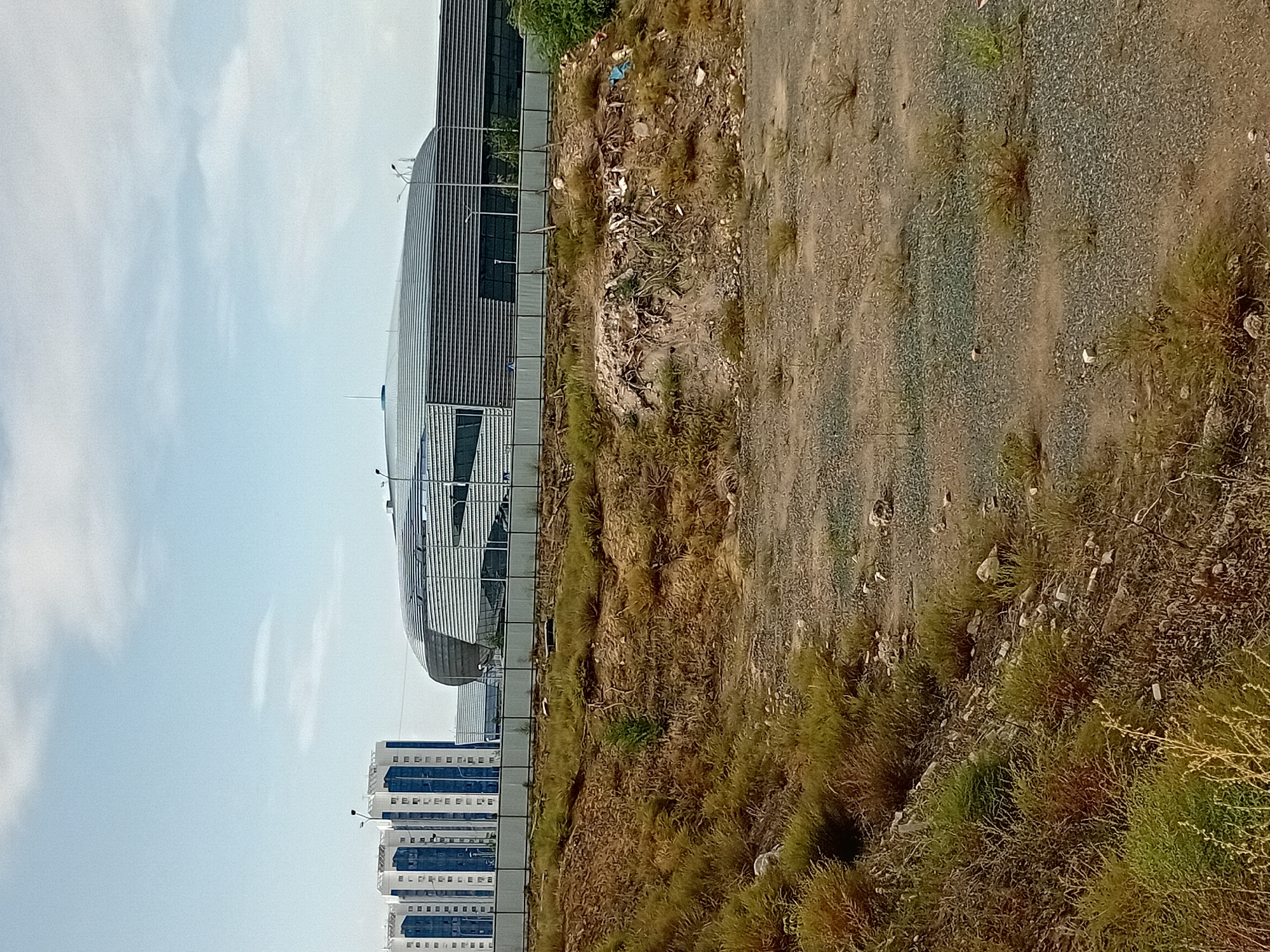 This is a landscape photograph taken in the city of Semey, Kazakhstan, depicting a modern stadium with a distinctive curved, metallic roof. In the background, several high-rise residential buildings with blue and white facades are visible. The foreground is an undeveloped, uneven terrain covered with sparse, dry vegetation and gravel, suggesting an area undergoing construction or left in a state of disrepair. A metal fence separates the undeveloped area from the stadium and residential buildings. The sky is partly cloudy with a pale blue hue. There are no people visible in the image. The overall impression is one of urban development juxtaposed with a neglected natural environment.