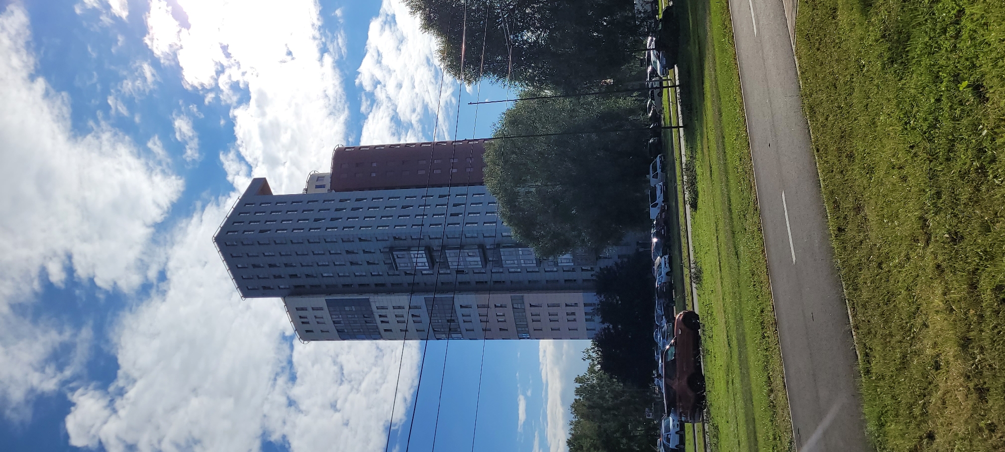 In this outdoor, daytime photograph taken in Riga, Latvia, a cluster of modern, high-rise apartment buildings dominates the background, set against a backdrop of a bright blue sky filled with white, fluffy clouds. The buildings, varying in height and architectural style, feature numerous windows and balconies, suggesting a residential area. A large, mature tree with dense green foliage stands in the middle ground, partially obscuring the buildings. In the foreground, a two-lane road runs horizontally across the image, with a grassy median separating it from a strip of manicured lawn. Several cars are parked along the side of the road in the middle ground, and one car, a reddish-orange SUV, is in motion on the road closest to the viewer. Overhead power lines crisscross the scene, adding a utilitarian element to the urban landscape. The overall impression is one of a contemporary urban environment, likely a residential district, under a pleasant sky.