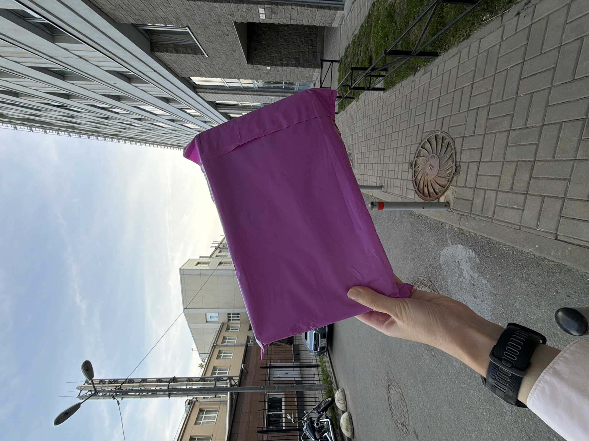 The image depicts a first-person perspective of an urban street scene in Saint Petersburg, Russia. In the immediate foreground, a light-skinned right hand, wearing a black sports watch with a textured band and prominent buckle on the left wrist, holds a crinkled, vibrant magenta/purple plastic mailer envelope. The sleeve of a light-colored shirt is visible on the lower forearm.
The scene unfolds along a paved sidewalk and an adjacent asphalt street. The sidewalk is constructed from grey interlocking concrete pavers laid in a herringbone pattern. Embedded in the sidewalk near the hand is a distinctive circular, ornate cast-iron manhole cover featuring a spiral or fan-like design. A low black metal railing separates the sidewalk from a narrow strip of grass. Two grey cylindrical bollards with red tops mark the edge of the sidewalk near the street.
To the left, on the asphalt street, a black cruiser-style motorcycle is parked beside several light-colored concrete spherical bollards. Further down the street, behind a black metal fence with vertical bars, a dark-colored sedan, resembling a Subaru WRX, is parked. The car's rear license plate is partially visible, displaying "178 RUS," which is the regional code for Saint Petersburg, Russia. Behind the fence, there is a light-colored metal gate and an older multi-story building with numerous windows. A tall, multi-faceted metal street lamp post with two luminaires and antenna attachments stands prominent on the left.
To the right, a large, modern multi-story building with a light grey facade and repetitive rows of windows dominates the background. An external ladder or fire escape is visible on its side. The ground floor of this building features a darker grey brick or stone facade with larger windows and a recessed entrance. Above the entrance, a subtle numeral "1" is visible.
The sky overhead is light blue with scattered wispy clouds, suggesting either early morning or late afternoon light.
