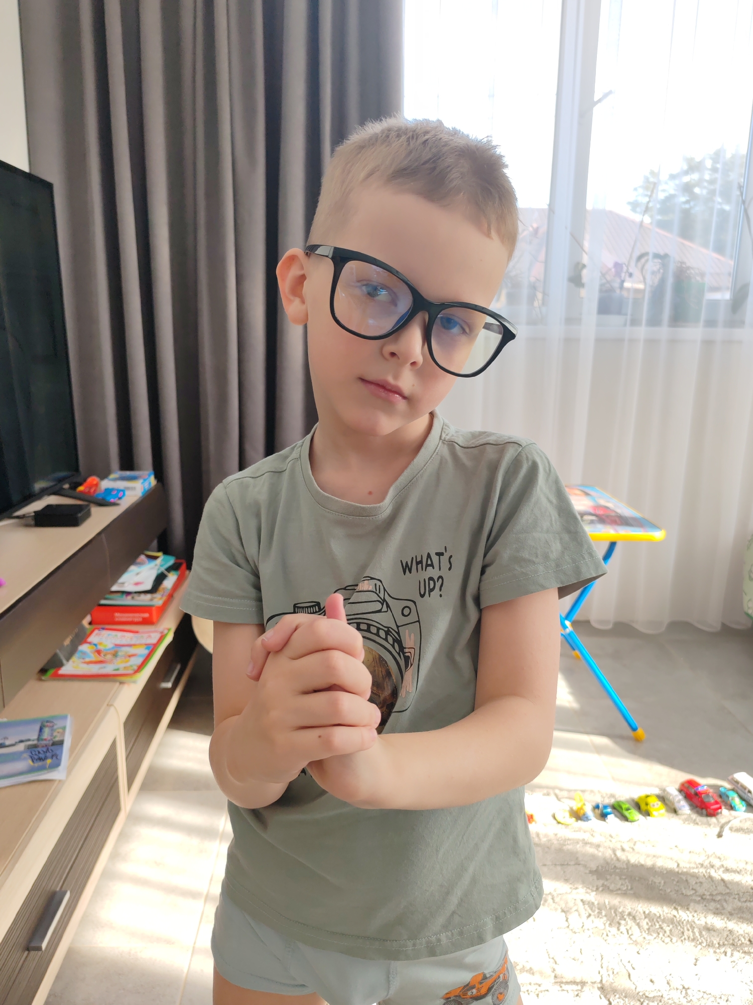 The image depicts an indoor residential scene in the city of Chaikovsky, Russia. A young male child, estimated to be between 5-7 years old, stands centered in the frame, directly facing the viewer with a neutral facial expression. He has short, light-colored hair and wears black-framed eyeglasses. He is dressed in a short-sleeved, sage green t-shirt with a graphic design of a camera and the text "WHAT'S UP?" on the front. He also wears light grey boxer briefs featuring an orange monster truck graphic on the left leg. His hands are clasped together at waist height.
The floor on the left side of the image consists of light-colored tiles, transitioning to a light-colored, shaggy area rug on the right. On the left, a multi-tiered wooden entertainment unit is visible, holding a partially obscured flat-screen television, a small black rectangular electronic device, and a remote control on its top surface. Below, multiple colorful children's books and magazines are stacked. On the rug to the child's right, a line of approximately eight small toy cars, including red, green, yellow, blue, and white vehicles, is arranged horizontally. In the background, a large window on the right is covered by sheer white curtains, offering a view of a light-tiled roof and green trees outside. A small child-sized table with a yellow surface and blue collapsible legs is partially visible behind the child on the right. Dark grey vertical curtains or blinds are drawn on the left side of the background. The scene is illuminated by bright natural light entering from the window, creating distinct shadows on the floor.