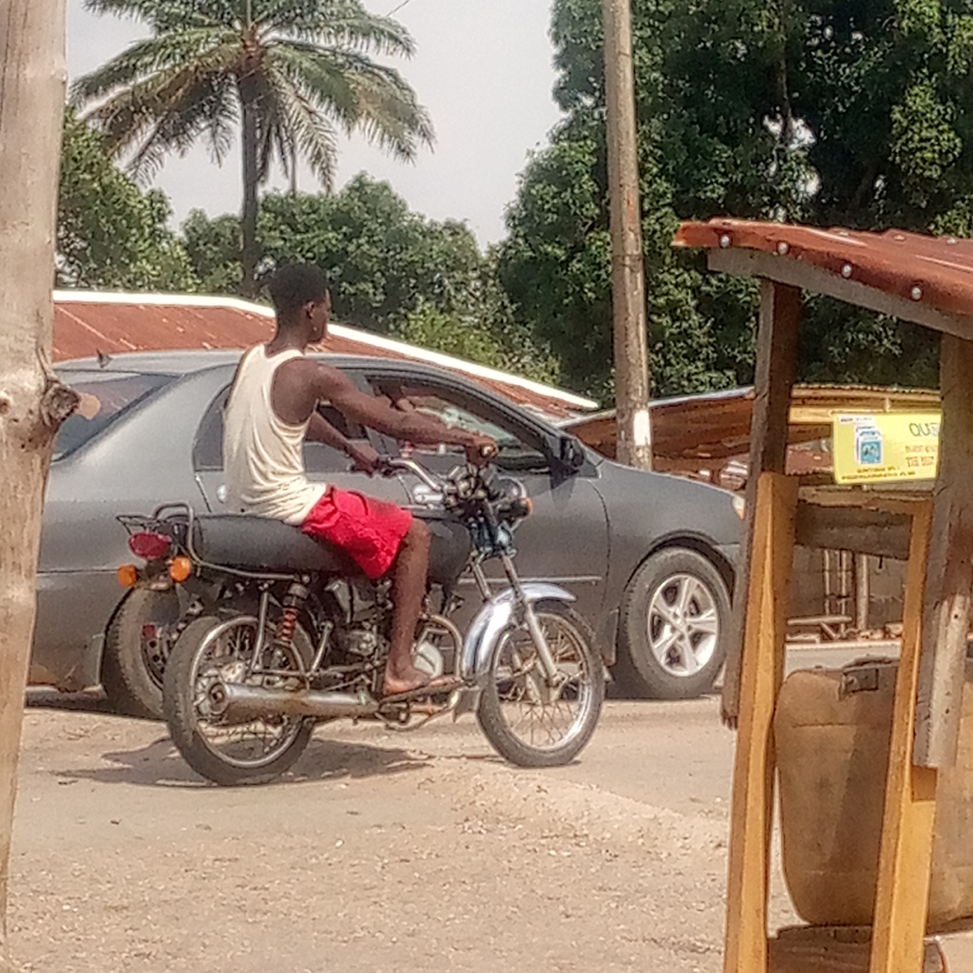 The scene is in Mararaba, Nigeria, showing a man on a motorcycle in a street environment. He wears a white tank top and red shorts, and rides barefoot. The motorcycle is black with chrome accents. A gray car is parked behind the motorcycle. In the background, there is a palm tree and other lush greenery. A makeshift wooden structure with a rusty metal roof is visible on the right side of the image. The road surface is dirt.