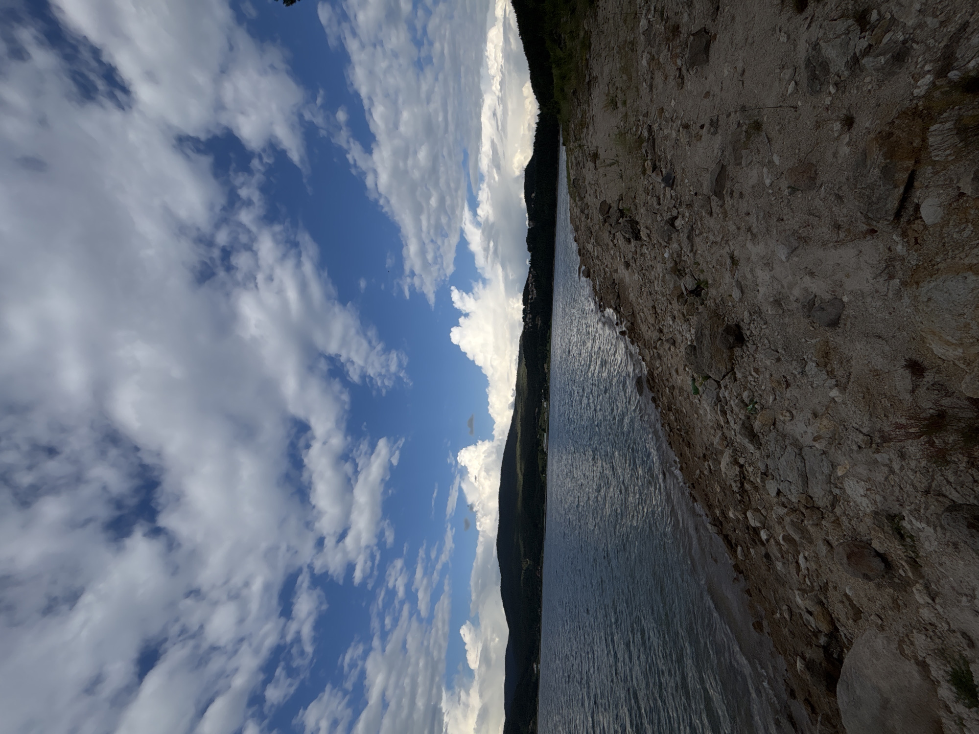 The image depicts a scenic natural landscape under a partly cloudy sky. The scene is dominated by a body of water, likely a lake or a large river, on the left side of the frame, reflecting the blue sky and white clouds. The water appears calm with gentle ripples. To the right of the water, a rocky shore is visible, with many stones and pebbles of varying sizes, suggesting a natural, undeveloped coastline. Beyond the shore, a densely forested hillside rises, creating a dark, verdant backdrop. The sky is a vibrant blue, dotted with fluffy white cumulus clouds that cast soft shadows. The lighting suggests it is daytime, possibly late afternoon, given the contrast and the shadows. There are no people or man-made objects clearly visible in the image, emphasizing the wild and serene nature of the setting. The overall mood is peaceful and tranquil, evoking a sense of being in nature. The image is oriented vertically, as if taken from a slightly elevated viewpoint looking down at the water's edge and across the landscape. No text is visible in the image.