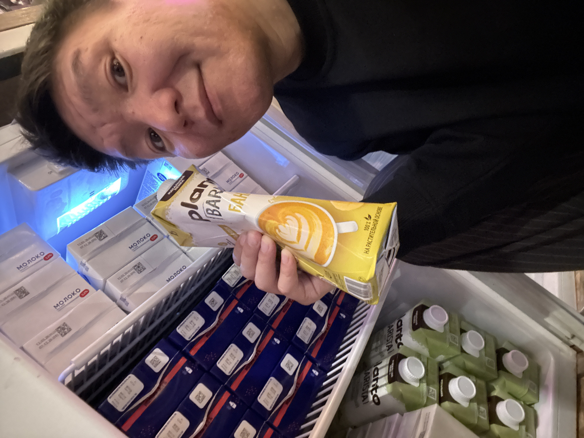 A man is taking a selfie in a refrigerator stocked with cartons of milk and plant-based drinks. The man is smiling and holding up a carton of what appears to be a plant-based coffee creamer with a picture of latte art on it. The refrigerator contains rows of white cartons labeled "МОЛОКО" (MILK) in Russian and green cartons with white caps. The image appears to have been taken inside a residential refrigerator, potentially in the city of Veshki, Russia, given the presence of Russian labeling.