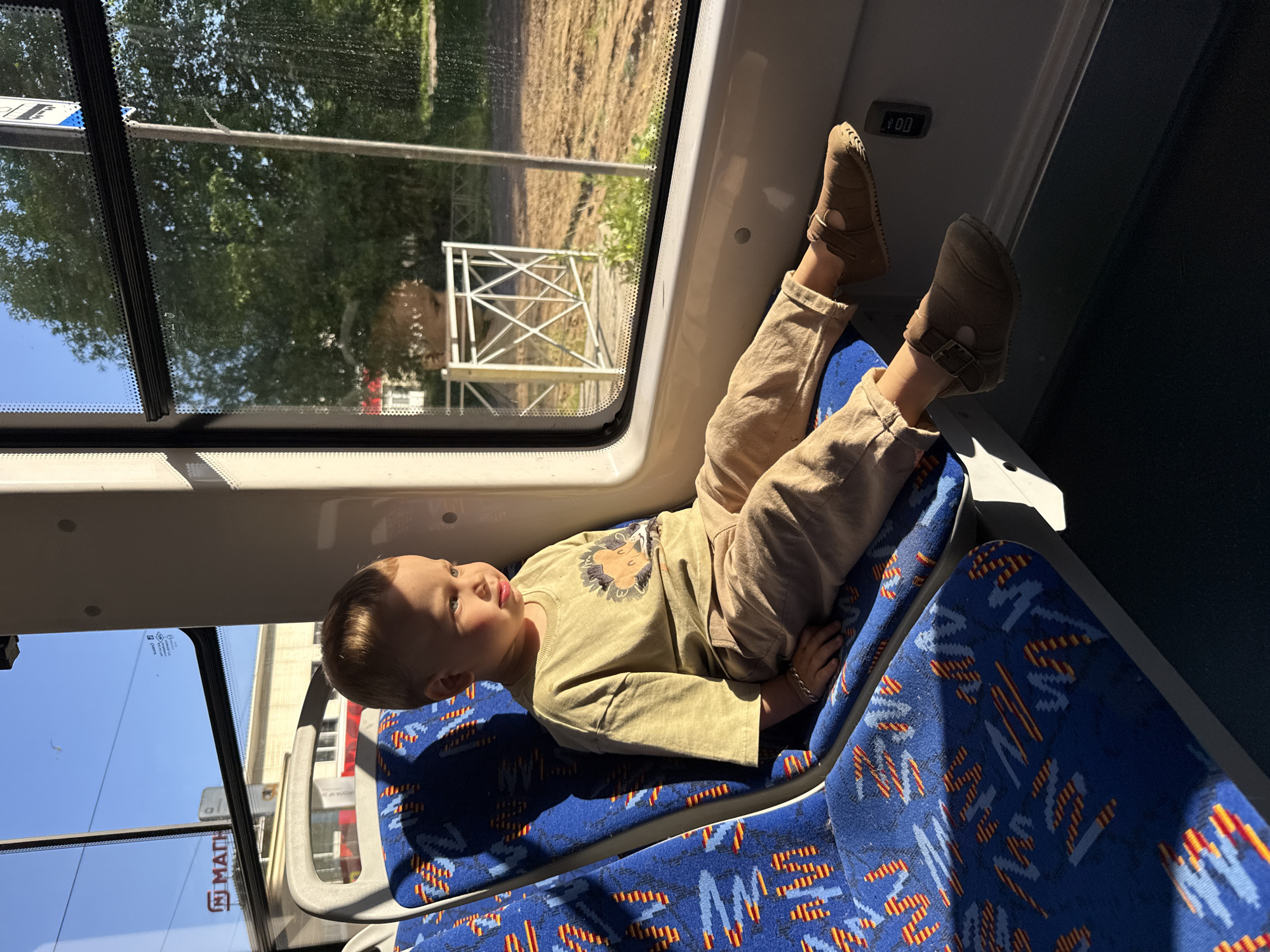 The image depicts the interior of a public transport vehicle, likely a tram or bus, in Rusanovo, Russia, during what appears to be either early morning or late afternoon due to the angle of direct sunlight. A young child, possibly a toddler or preschooler with light-colored hair and fair skin, is seated on a blue fabric seat with a pattern of red, orange, and light blue abstract zigzags. The child is wearing an olive-green t-shirt featuring a cartoon lion or hedgehog graphic, light brown/khaki trousers, and dark brown buckled shoes. A silver-colored bracelet is visible on the child's left wrist.
The child is positioned facing slightly towards the right-side window, with legs extended across the adjacent seat. Strong sunlight illuminates the child's right side, casting a distinct shadow on the seat to the left. The interior features light grey or white panels and window frames. A small black rectangular device is affixed to the panel below the right window.
Through the windows, elements of the exterior are visible. On the left side, beyond the child's position, a clear blue sky is seen above distant buildings, one with a red roof. A sign with the Cyrillic letters "МАТН" in red is visible on a building or structure. Through the right-side window, dense green foliage and trees dominate the mid-ground. Below the trees, the ground appears dry and light brown, possibly unpaved or a dirt path. A white metal fence with a criss-cross design is discernible in front of the foliage, and a thin grey pole stands vertically. Faint reflections of the child's head are visible on the window glass, along with minor smudges or droplets.
