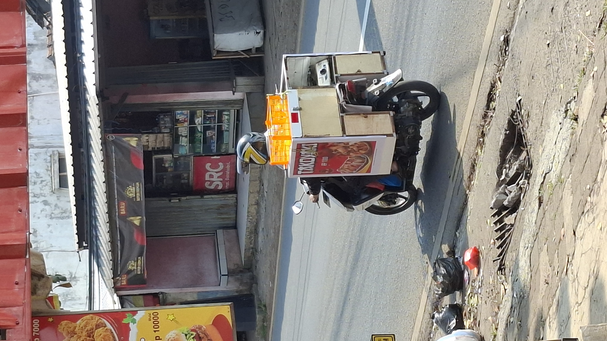 A person rides a motorcycle with a food cart attached to the back down a street in Cilaku, Indonesia. The food cart is white with wooden accents and displays the text "TAKOYAKI" prominently. It also has an orange basket on top. The motorcycle is black and white, and the rider is wearing a helmet. In the background, there is a store with a banner that says "BARU" (new) and "SRC". The street appears to be paved, and there is a storm drain on the side of the road with some debris in it. The scene is illuminated by daylight.