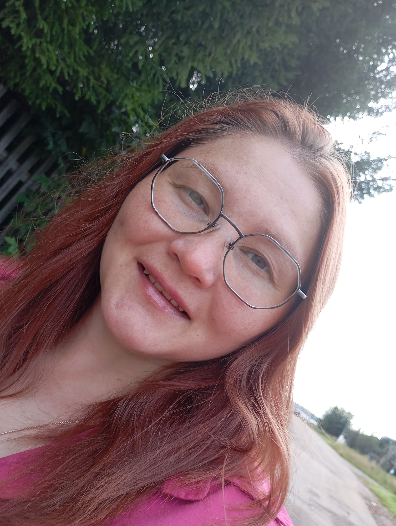 A medium shot, outdoor, eye-level, selfie shows a light-skinned woman with auburn hair and freckles. She is wearing glasses with thin, hexagonal frames and a bright pink top. She is smiling with her mouth slightly open, revealing her teeth. Her hair is long and wavy, and a portion of her neck and a thin rose gold necklace are visible. The background is a blur of green foliage and a bright, overcast sky, with a hint of a fence on the left and a road and trees in the distance on the right. The scene appears to be a residential area, possibly in the city of Fokino, Russia, given the presence of a fence, road, and trees.